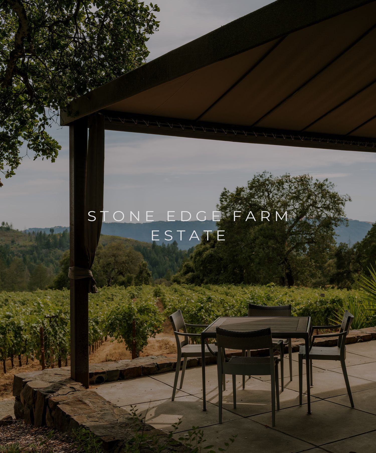 A covered patio with seating overlooks Stone Edge Farm Estate's scenic vineyard and rolling hills in the background.