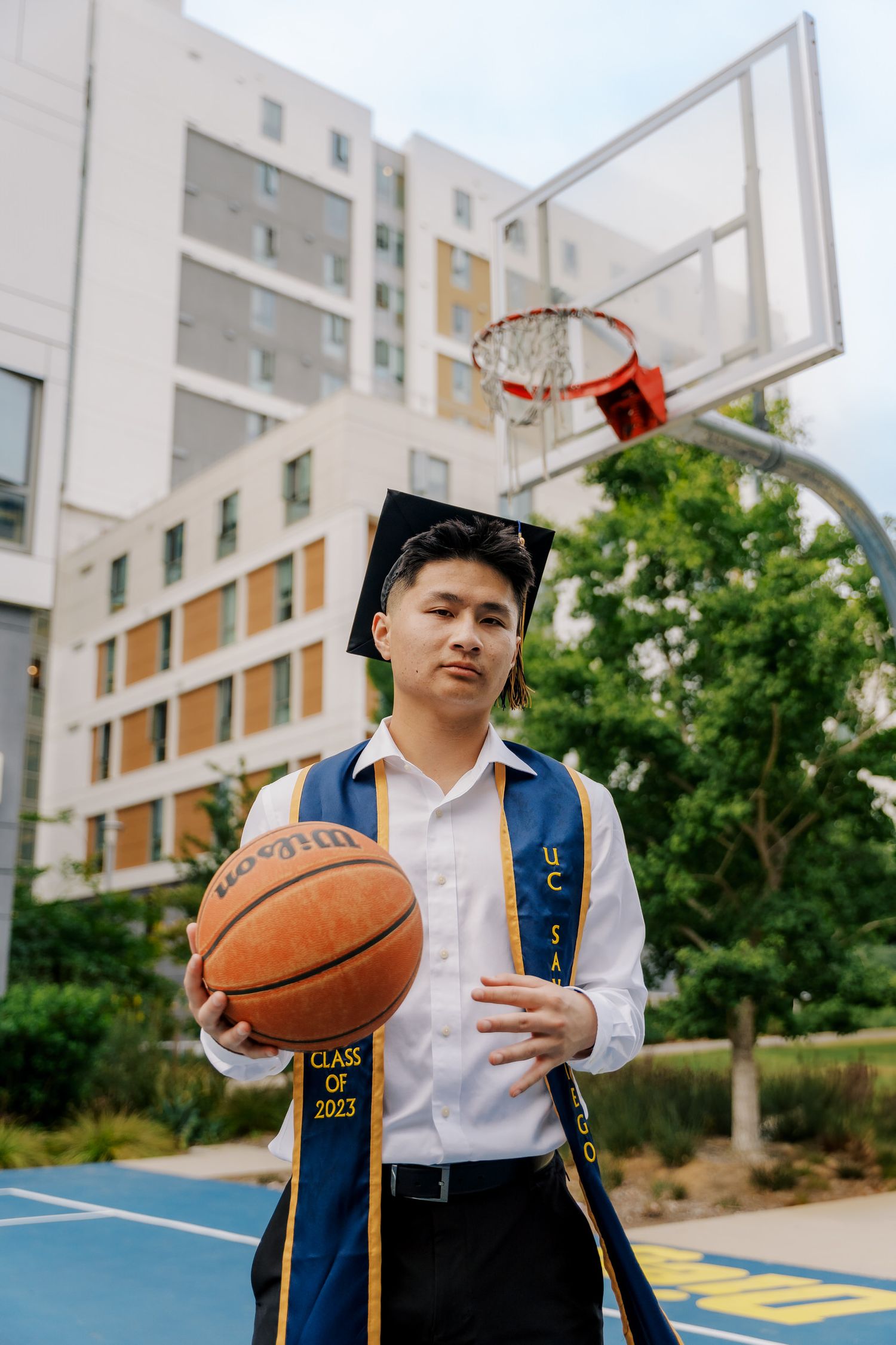 Graduate in academic regalia holds basketball on outdoor court with tall building in background.