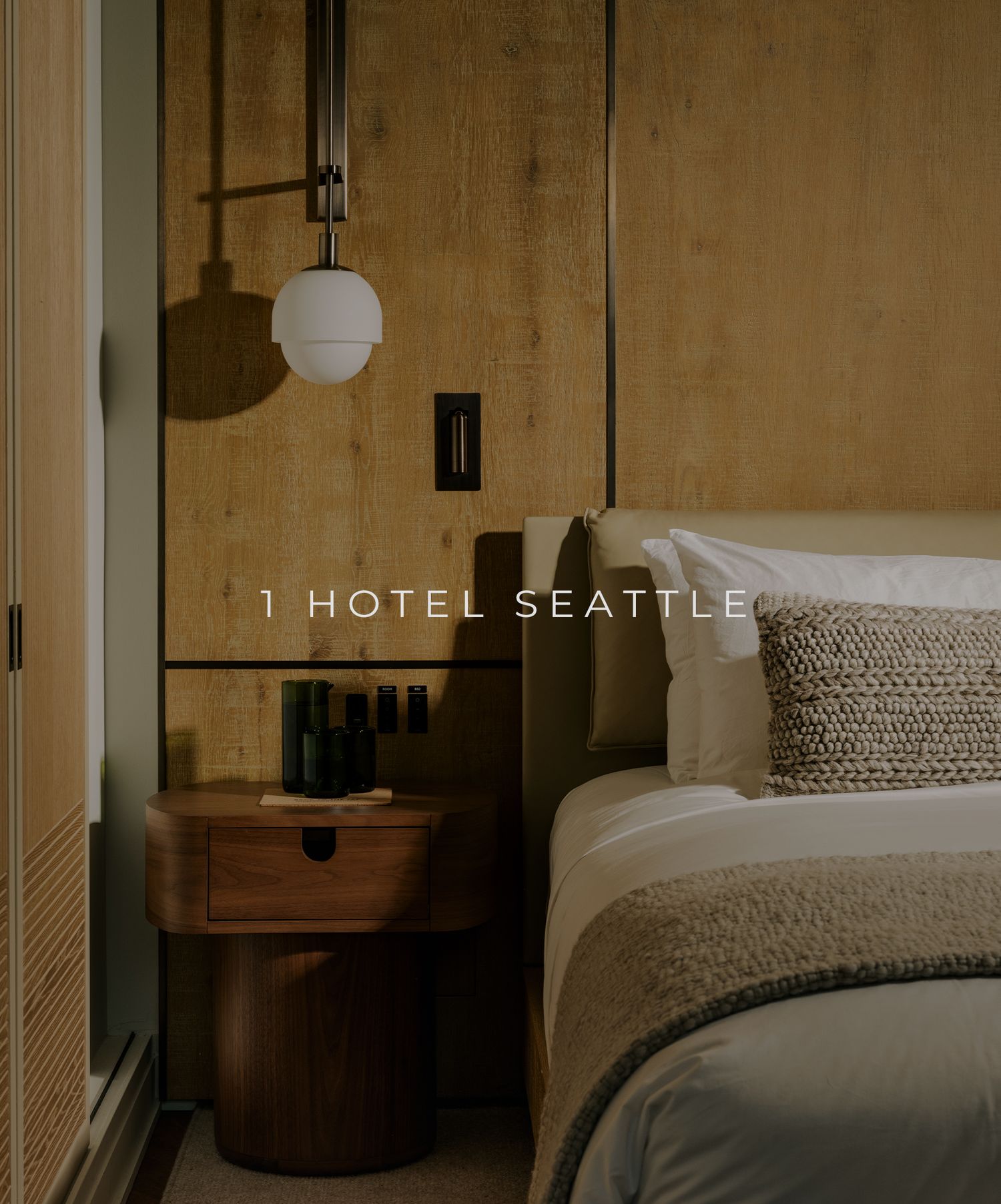 A modern hotel room interior with warm lighting, wood side table, globe pendant lamp and textured bedding against beige walls.