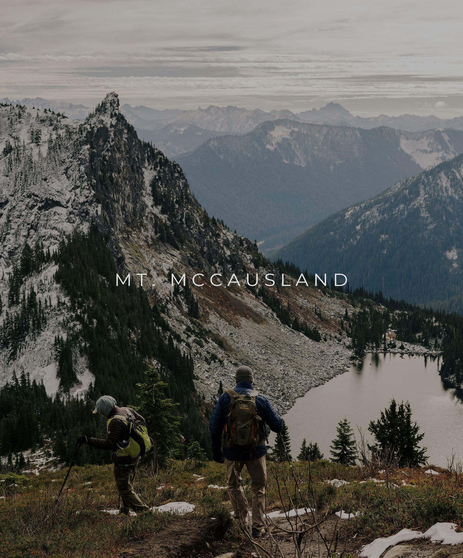 Two hikers stand at Mount McCausland summit overlooking a mountain lake and dramatic peaks in the Pacific Northwest wilderness.