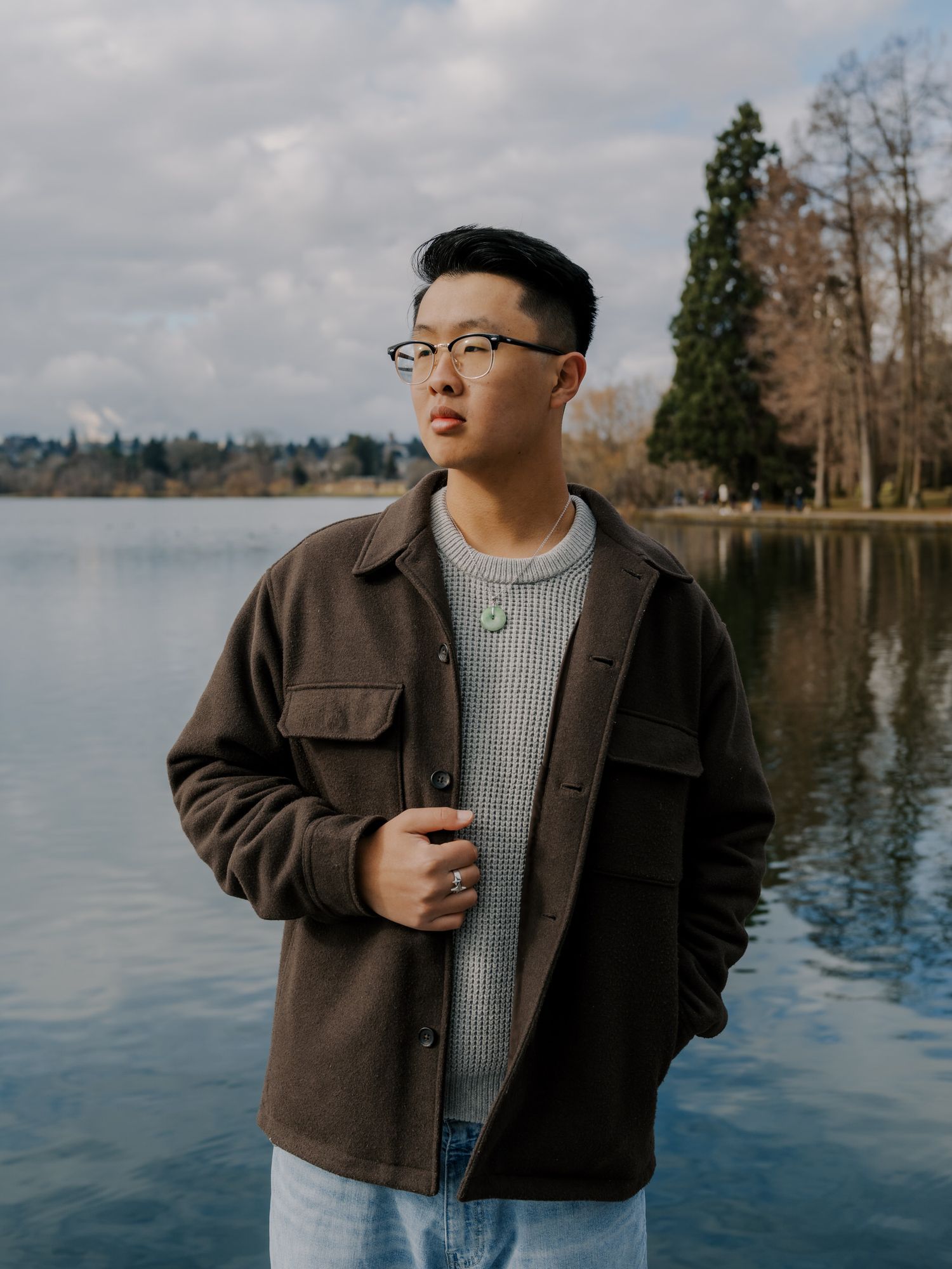 Person in glasses and brown jacket stands by tranquil lake with trees in background on overcast day.