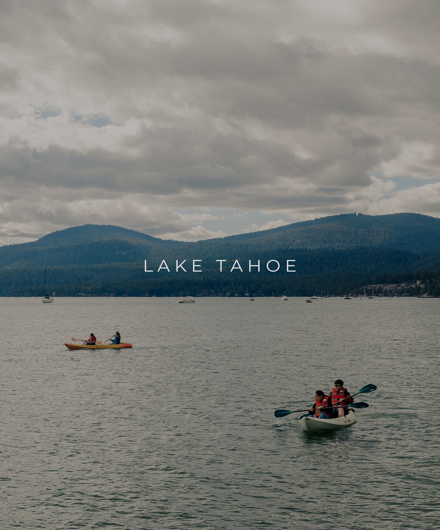 Kayakers paddle on Lake Tahoe's calm waters under cloudy skies with mountain ranges in the background.
