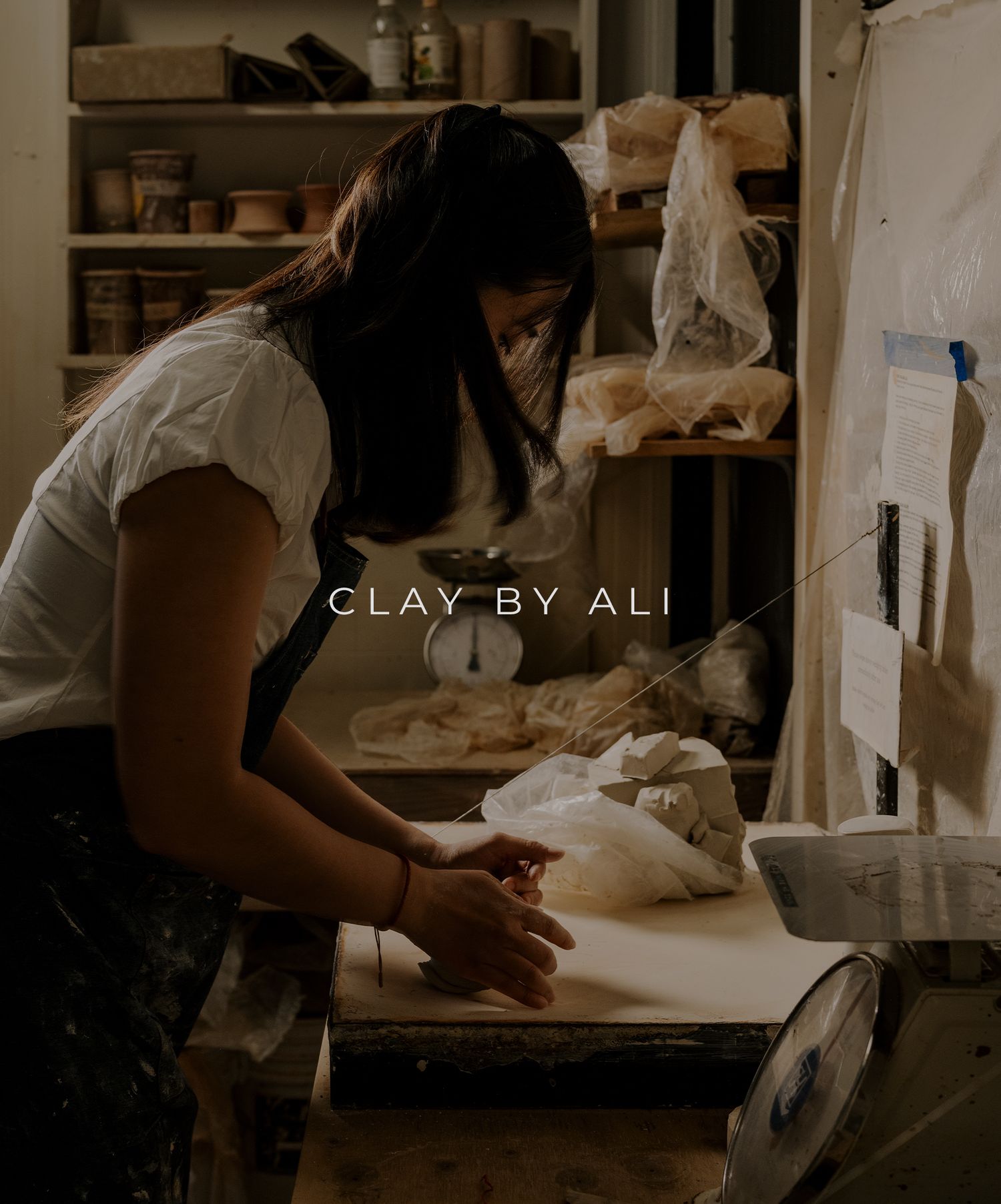 An artisan works at a pottery studio workbench with clay and tools visible on shelves in warm lighting.