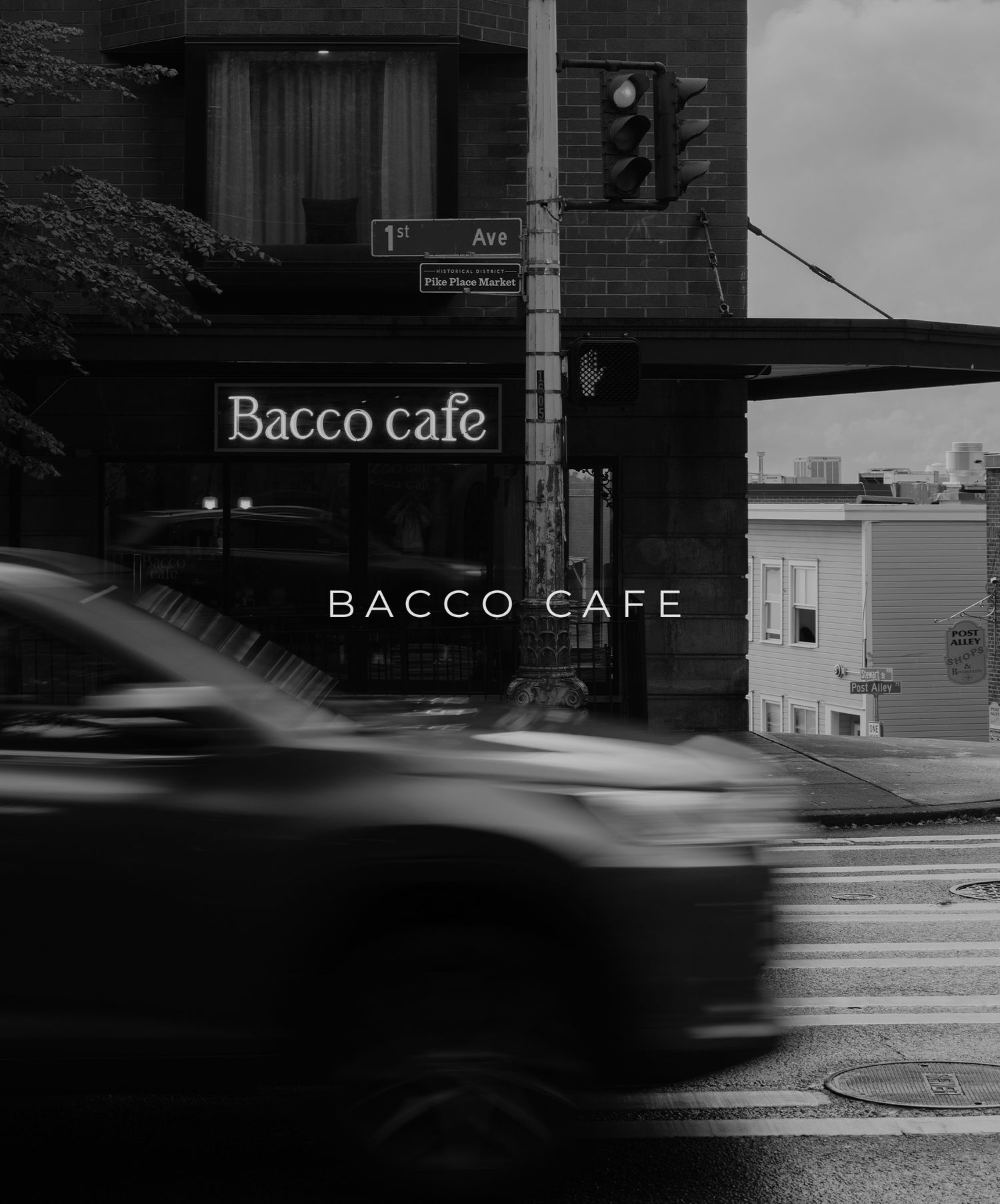 A blurred car passes by Bacco Cafe on a rainy street corner at dusk with illuminated signage in black and white.