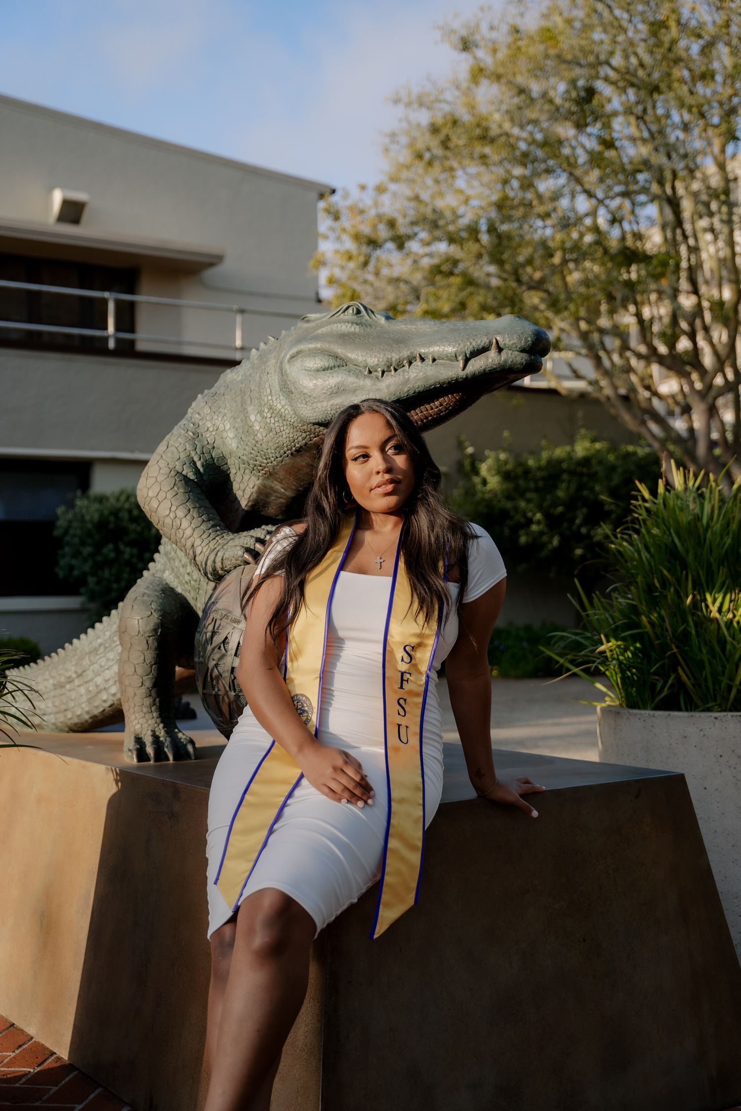 Graduate in white dress and gold sash poses with decorative alligator statue on campus.