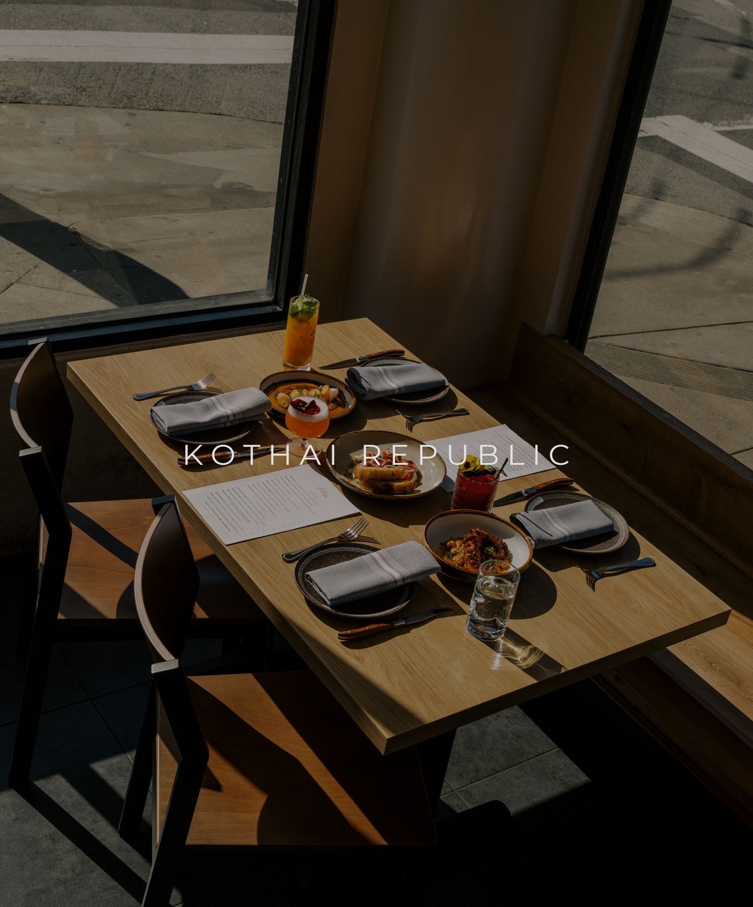Modern restaurant interior with wooden tables and place settings arranged in a dimly lit dining space.