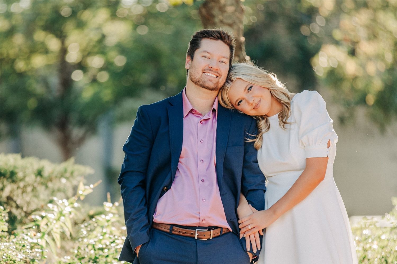 A couple stands close together in an outdoor setting with natural sunlit foliage in the background.