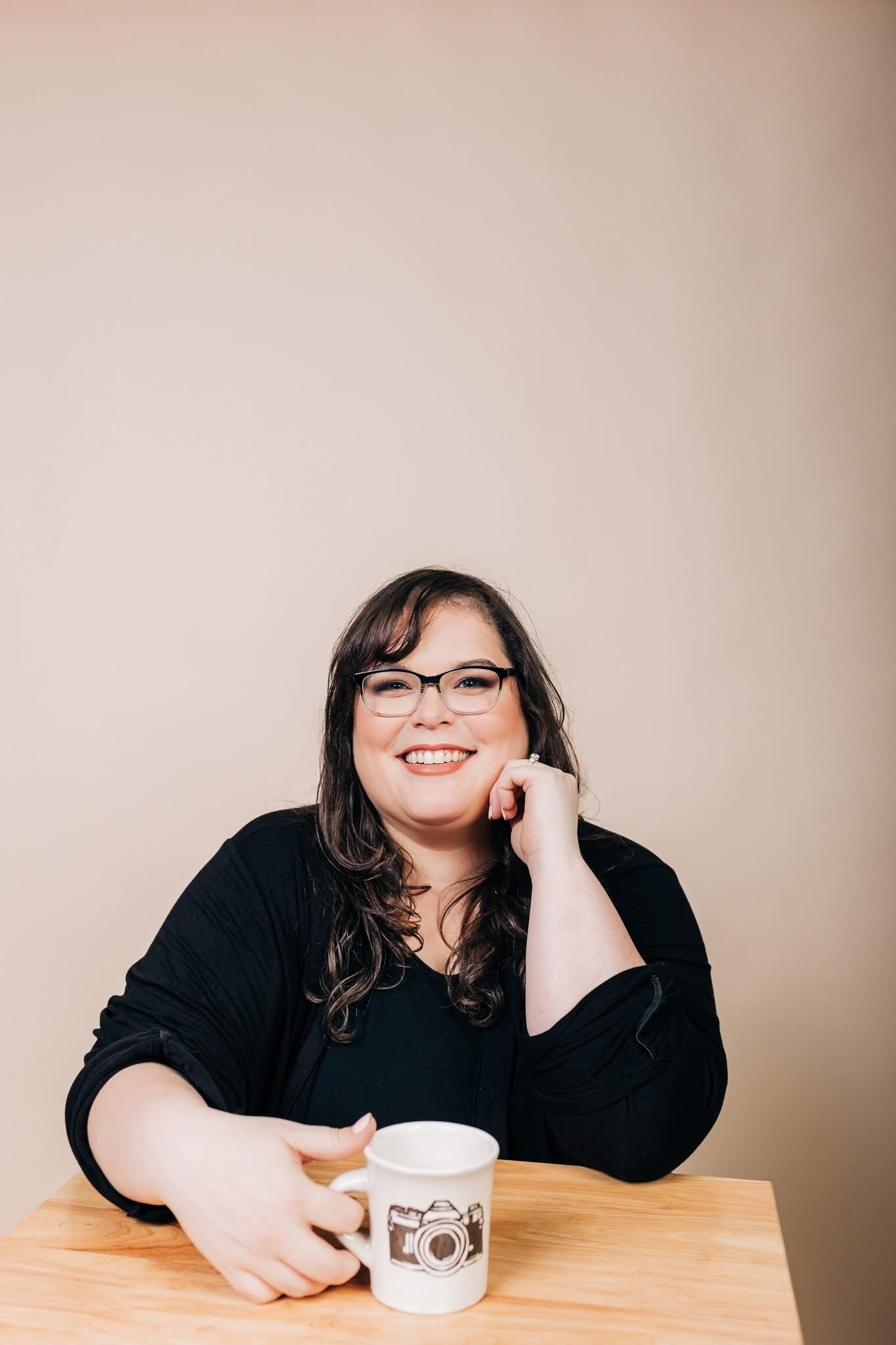Someone in a black sweater and glasses sits at a wooden table with a coffee mug against a beige background.