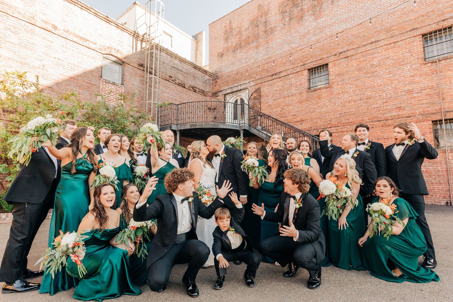 Wedding party in emerald green dresses and black tuxedos pose together against brick wall outdoors.
