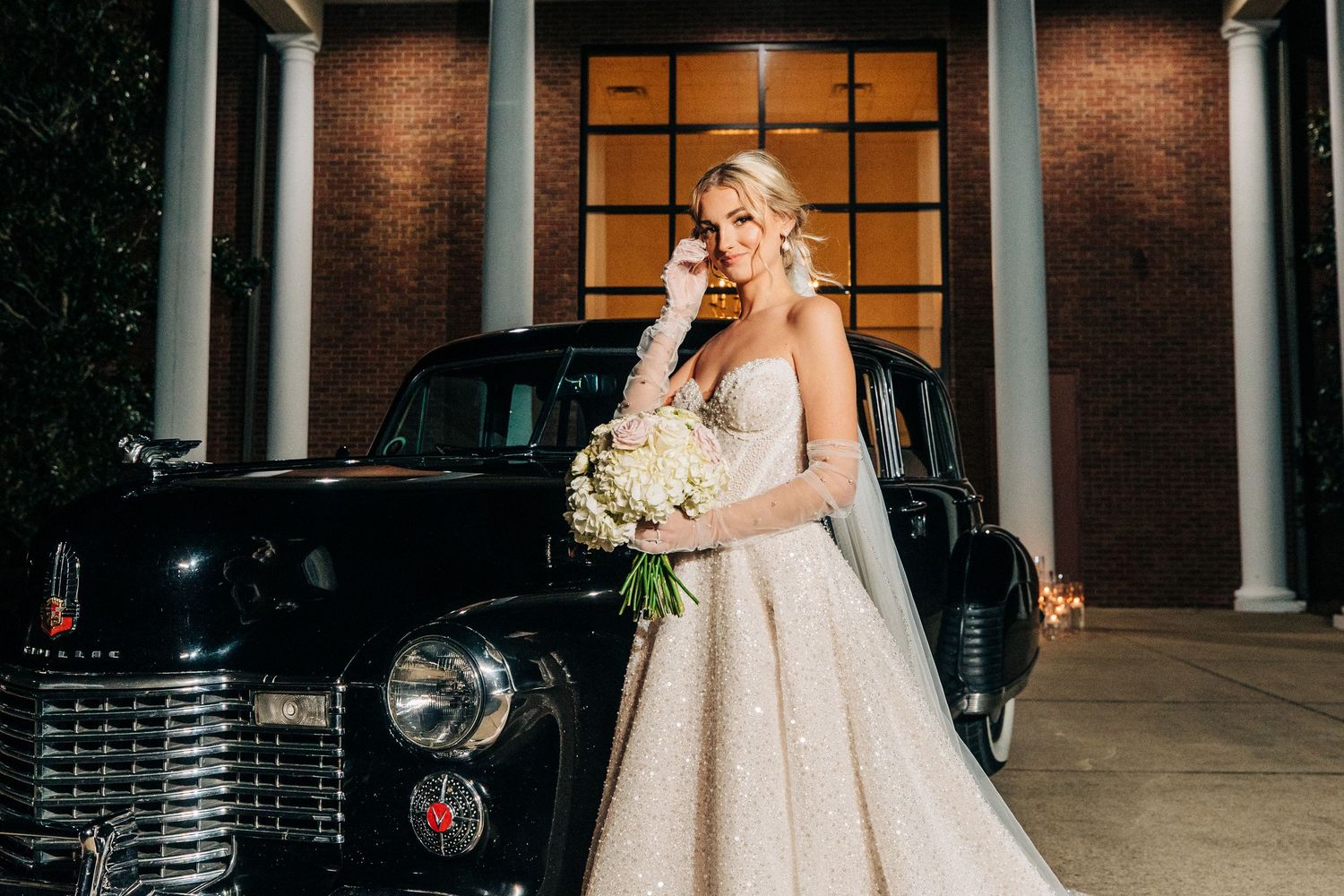 A bride in a sparkly white ballgown poses next to a classic black vintage car while holding a bouquet of flowers.