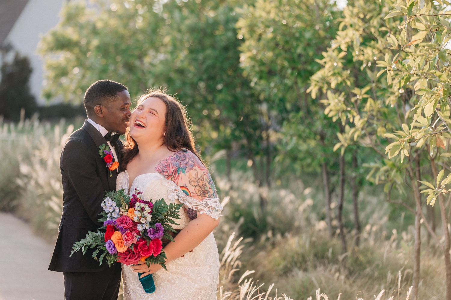 A joyful wedding couple shares an intimate moment outdoors while holding a vibrant red and pink floral bouquet.