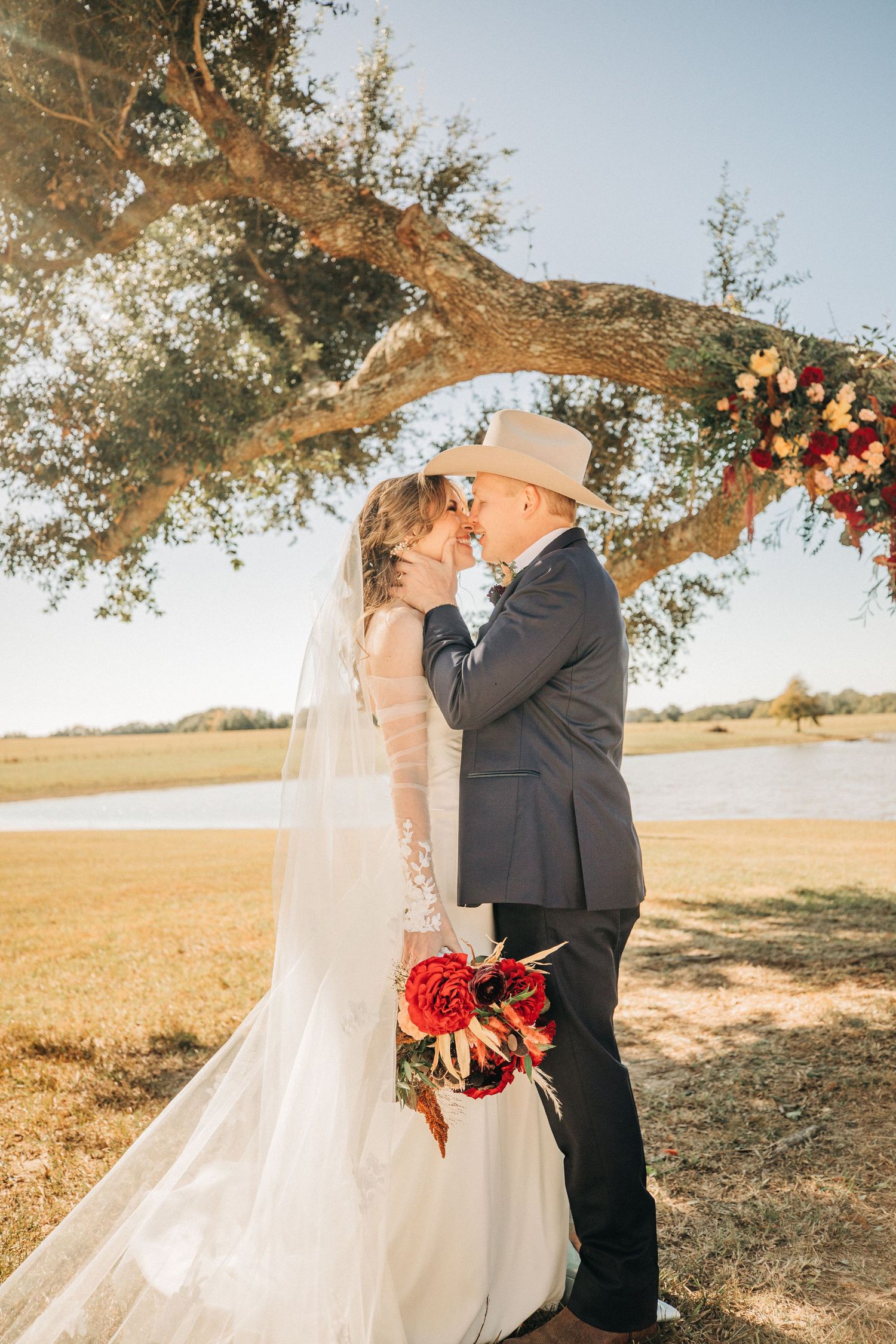 A romantic wedding moment captured under a sunlit tree branch decorated with floral arrangements and trailing veil.