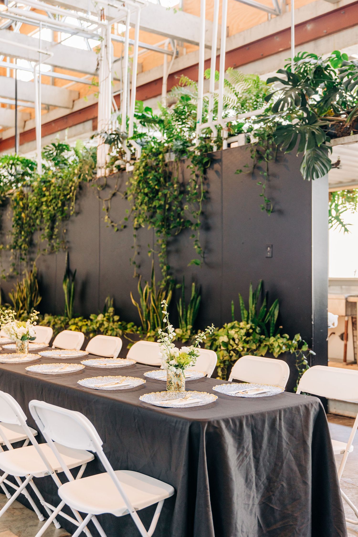 Elegant indoor dining setup with black tablecloths and white chairs surrounded by potted plants and hanging greenery.
