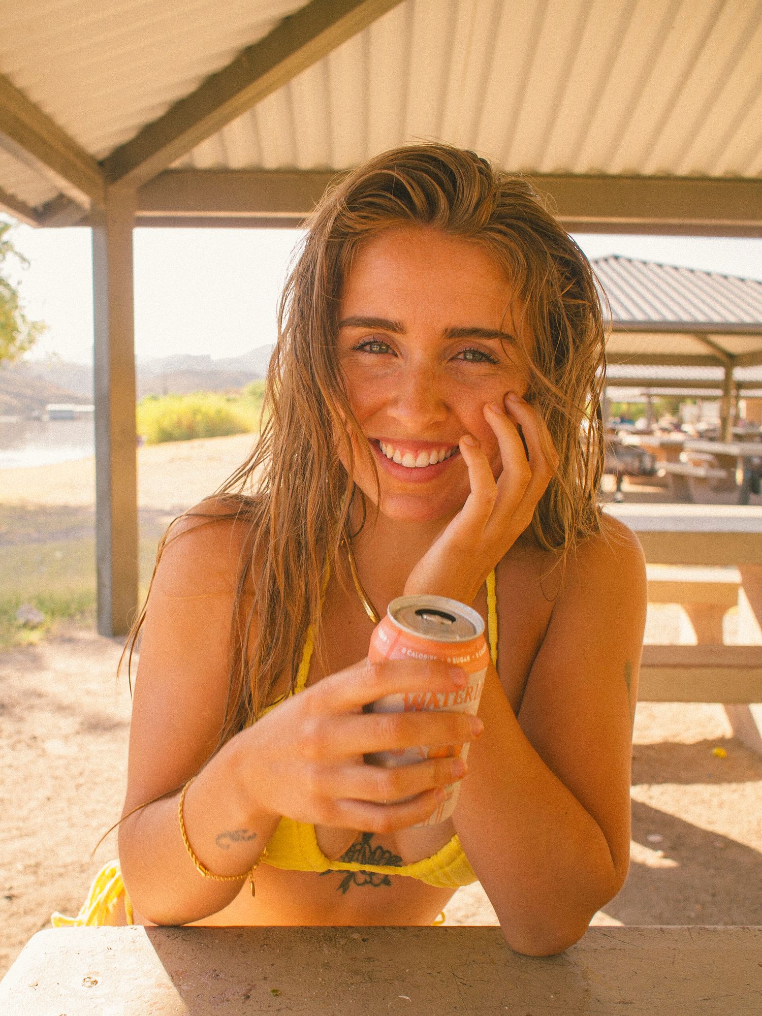 Young woman in yellow swimwear enjoying a sparkling water at an outdoor picnic table at the lake.