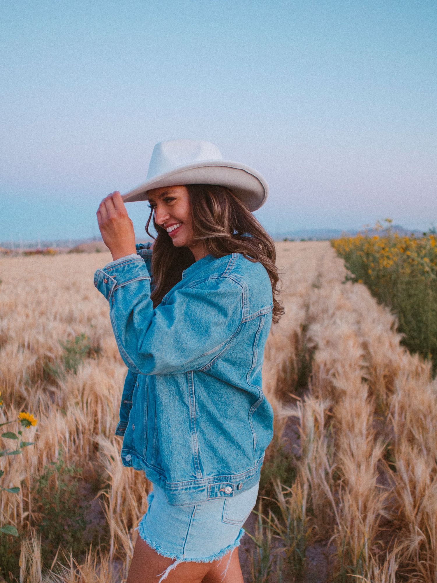 Young woman in denim jacket and white cowboy hat laughs while standing in golden wheat field under soft blue sky.