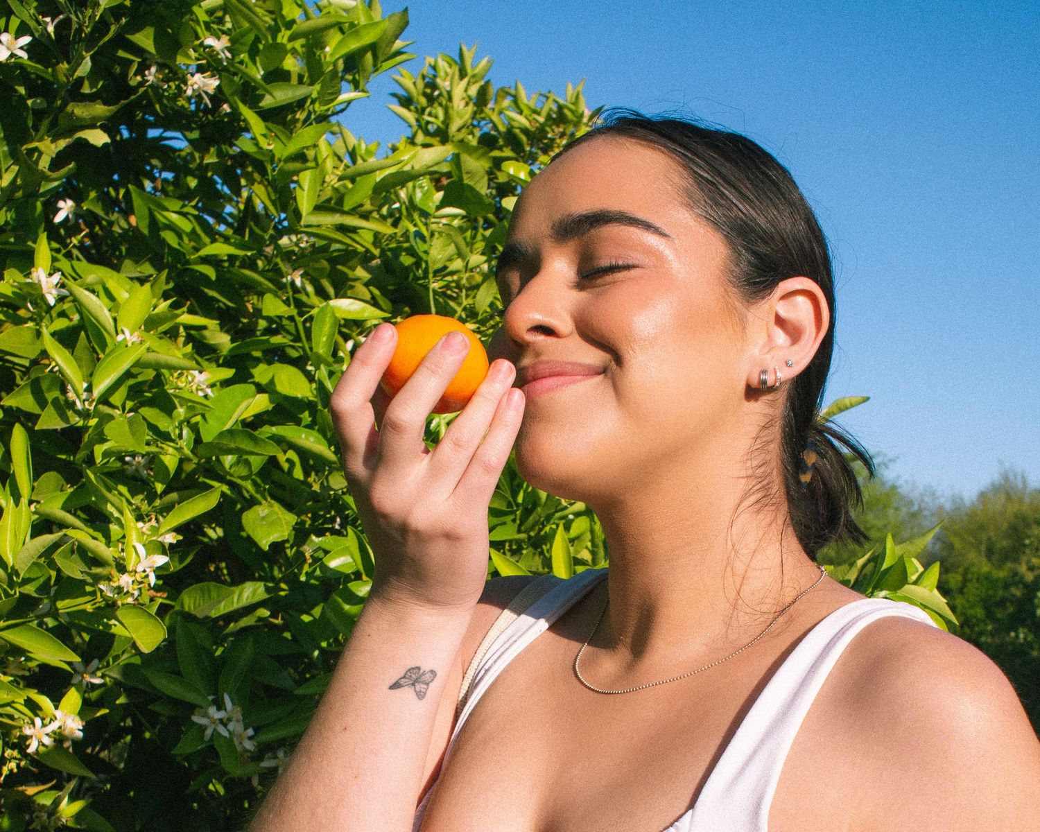 A woman in a white tank top smells a fresh citrus fruit while standing in front of green citrus trees on a sunny day.