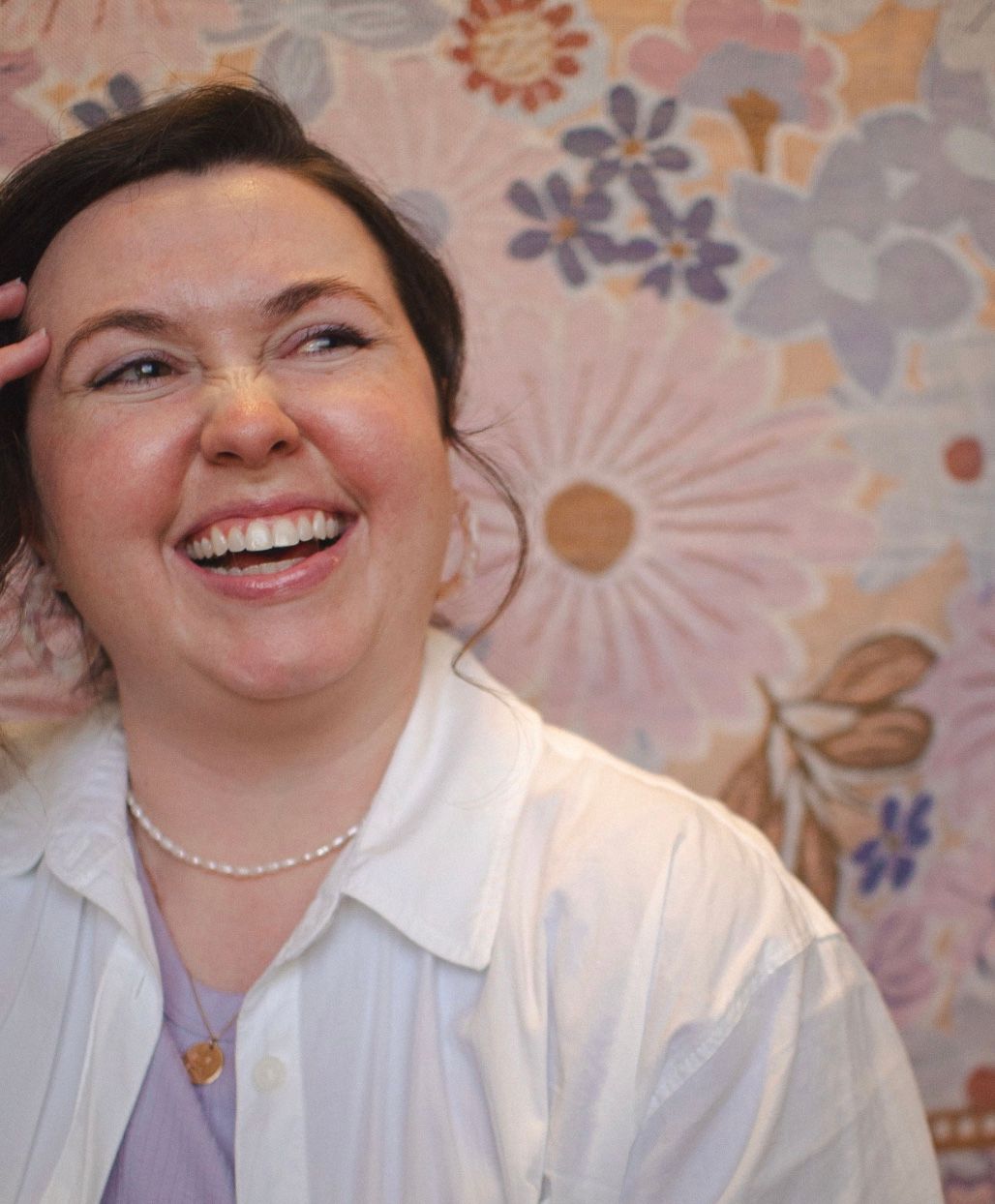 Kelsey, in a white collared shirt and pearl necklace, laughs joyfully against a floral blanket background.