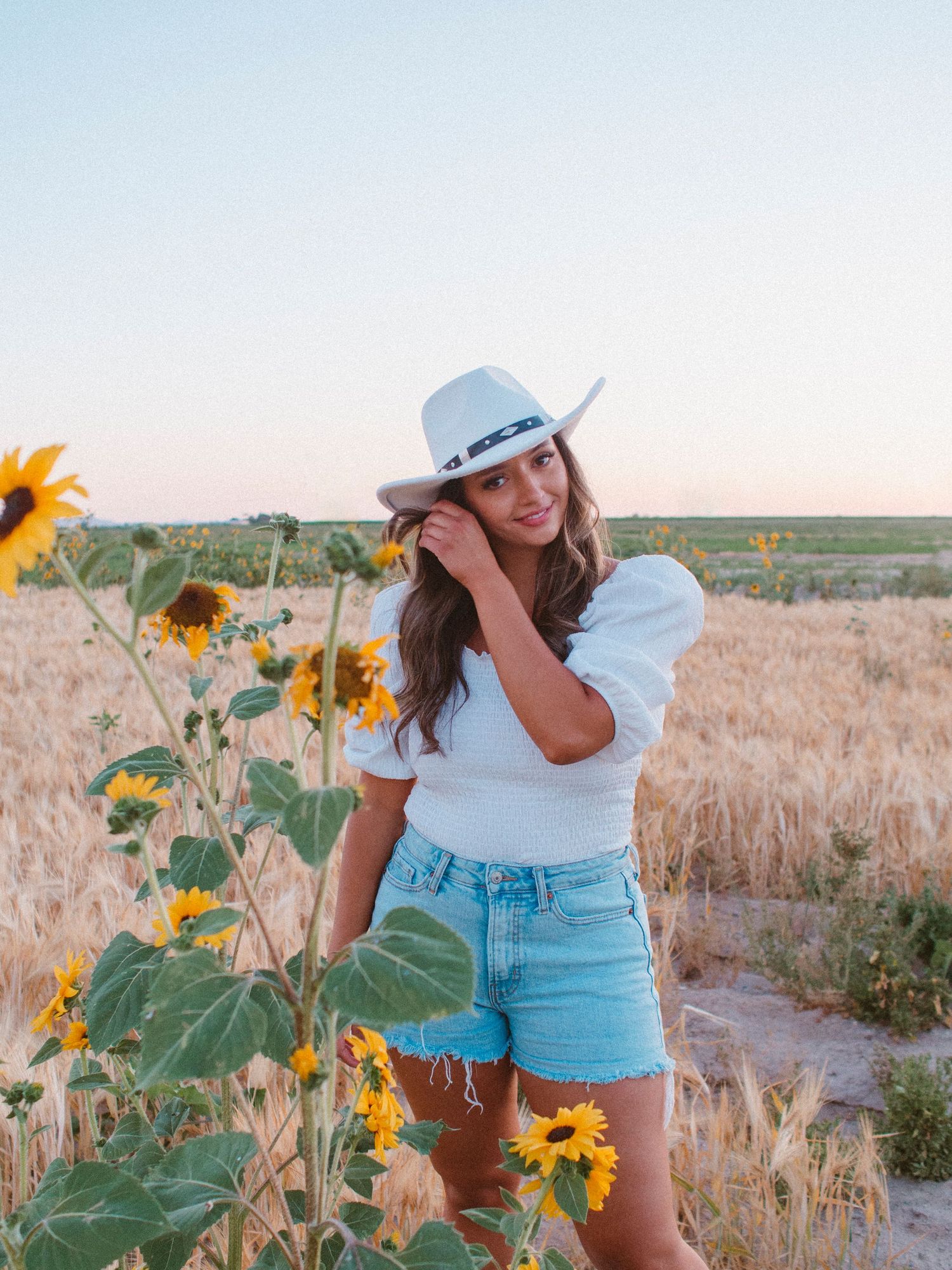 A woman in denim shorts and white top poses in a field of sunflowers during sunset while wearing a cowboy hat.
