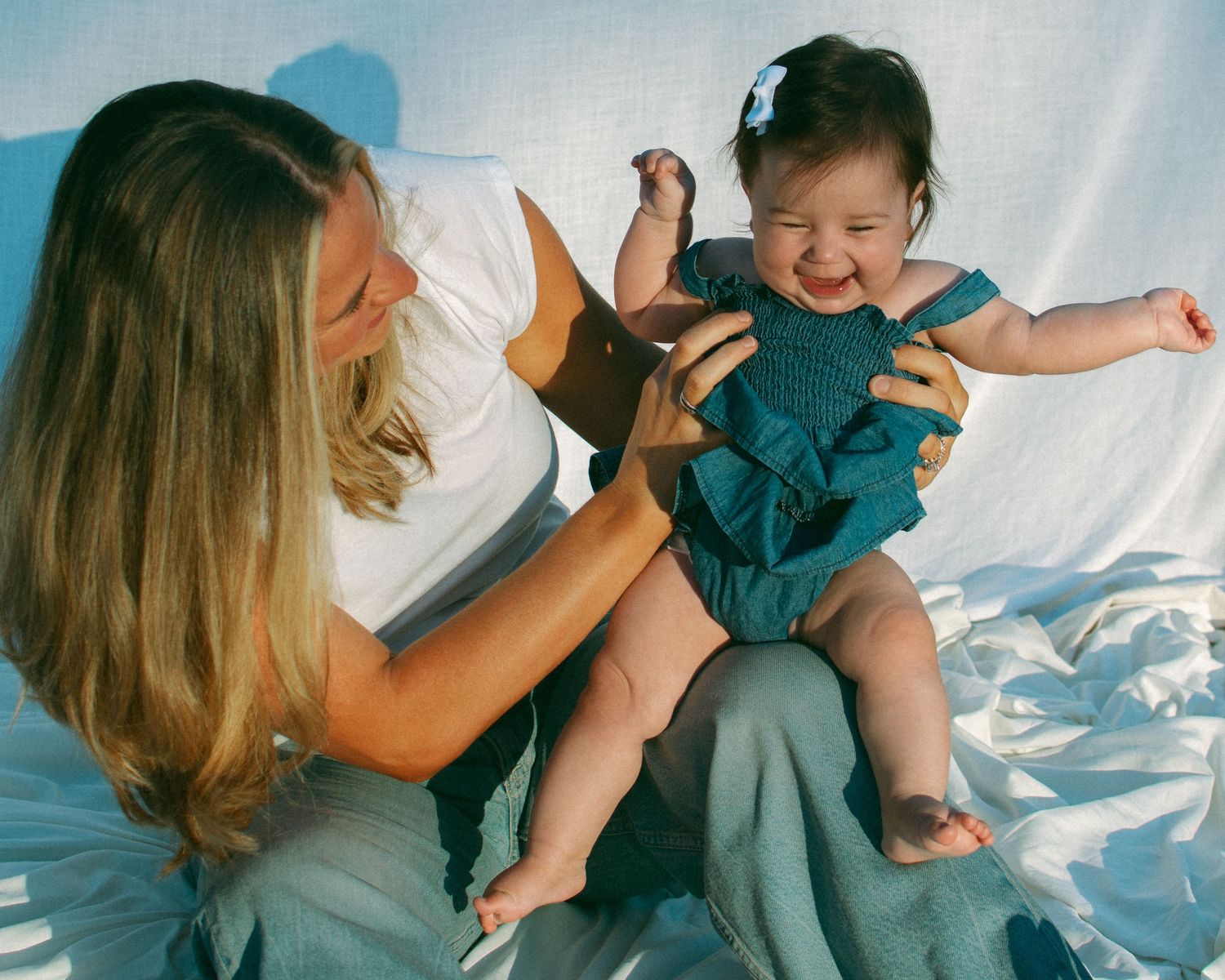 A joyful baby wearing a sweet denim outfit laughs while being playfully held by her mom on a white sheet backdrop.