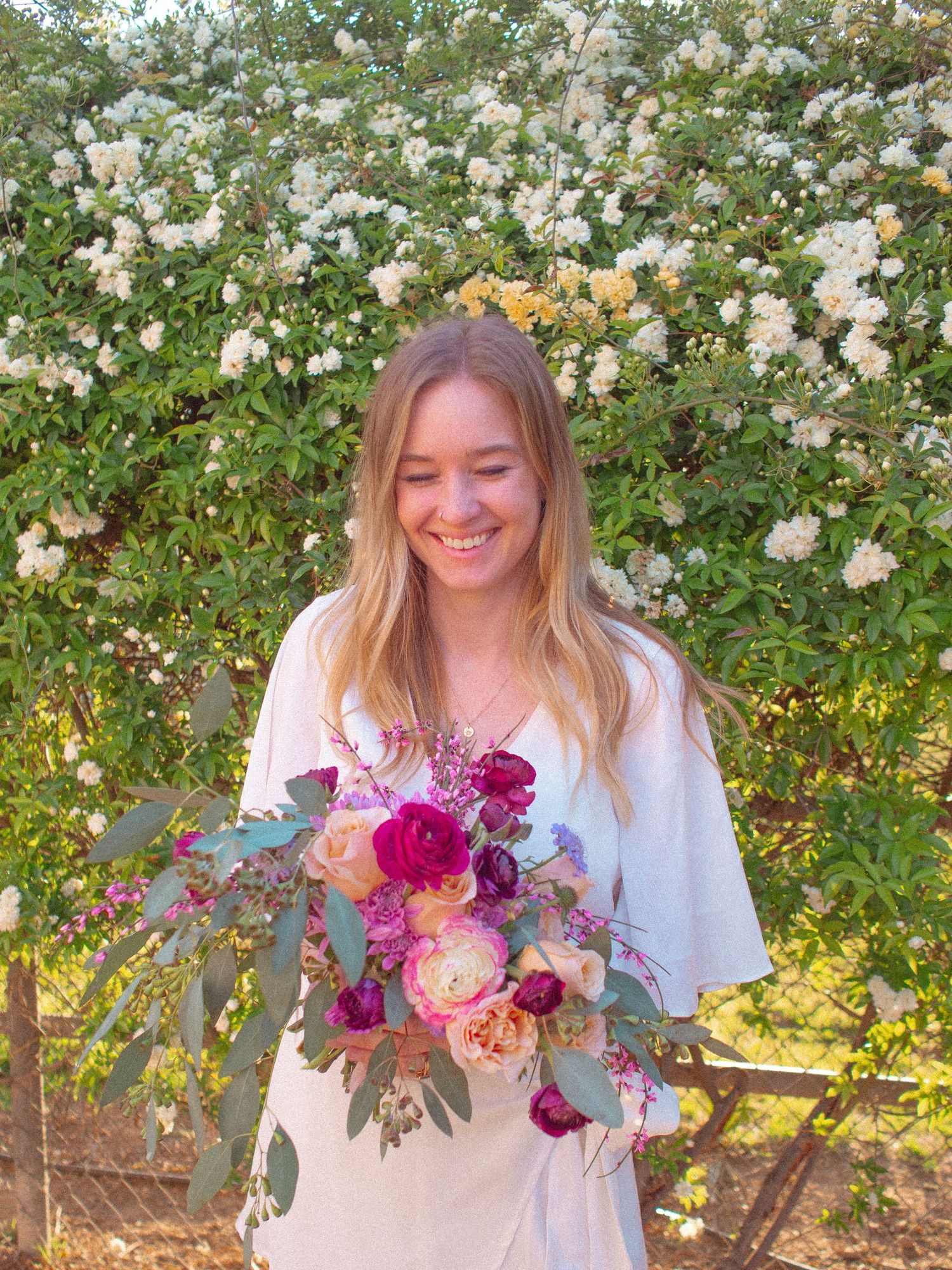 A cheerful young woman in a white dress holds a vibrant bouquet of pink and purple flowers against a backdrop of blooming roses.