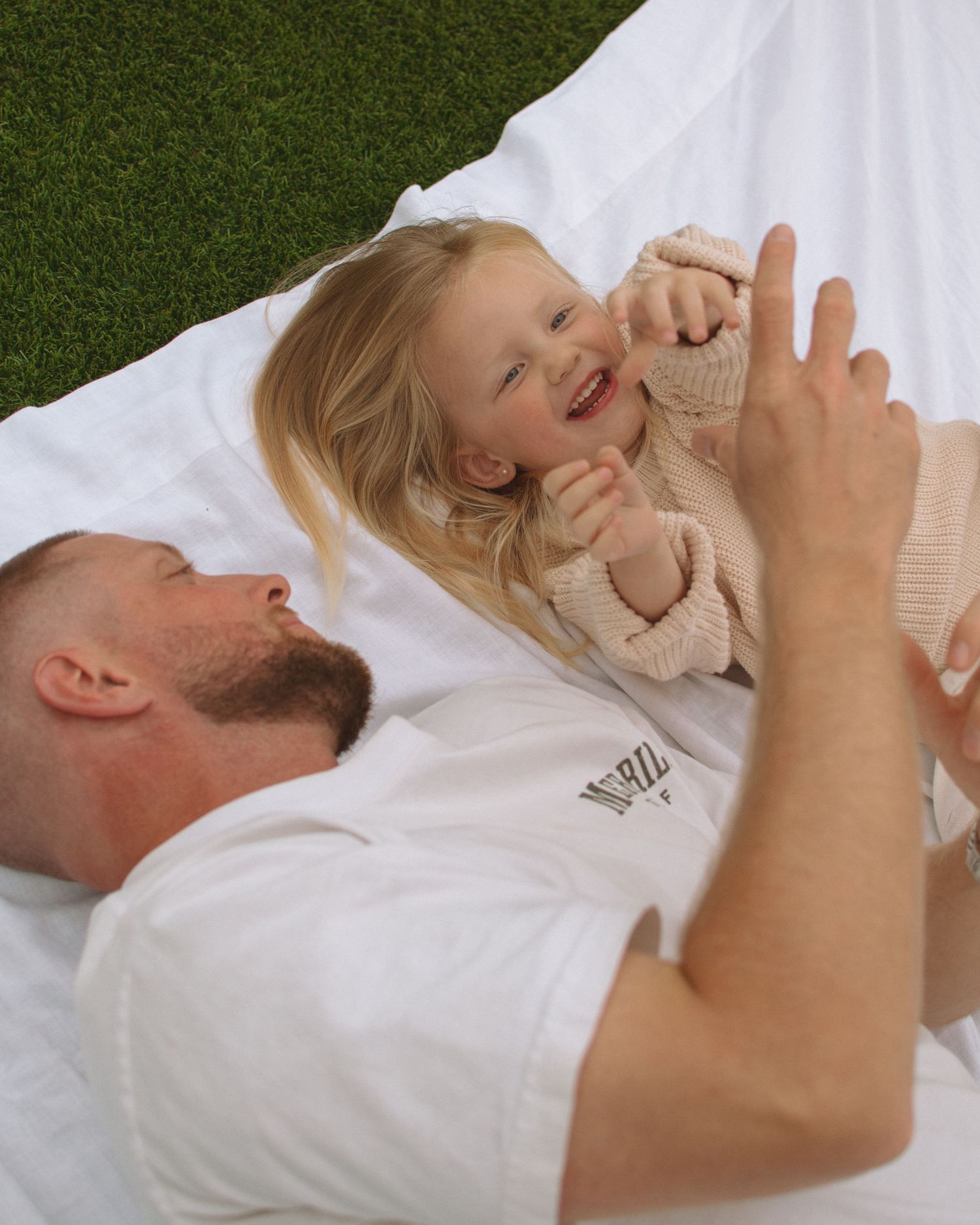 A parent and child share a joyful moment laying on a white blanket over grass, playing and laughing together.