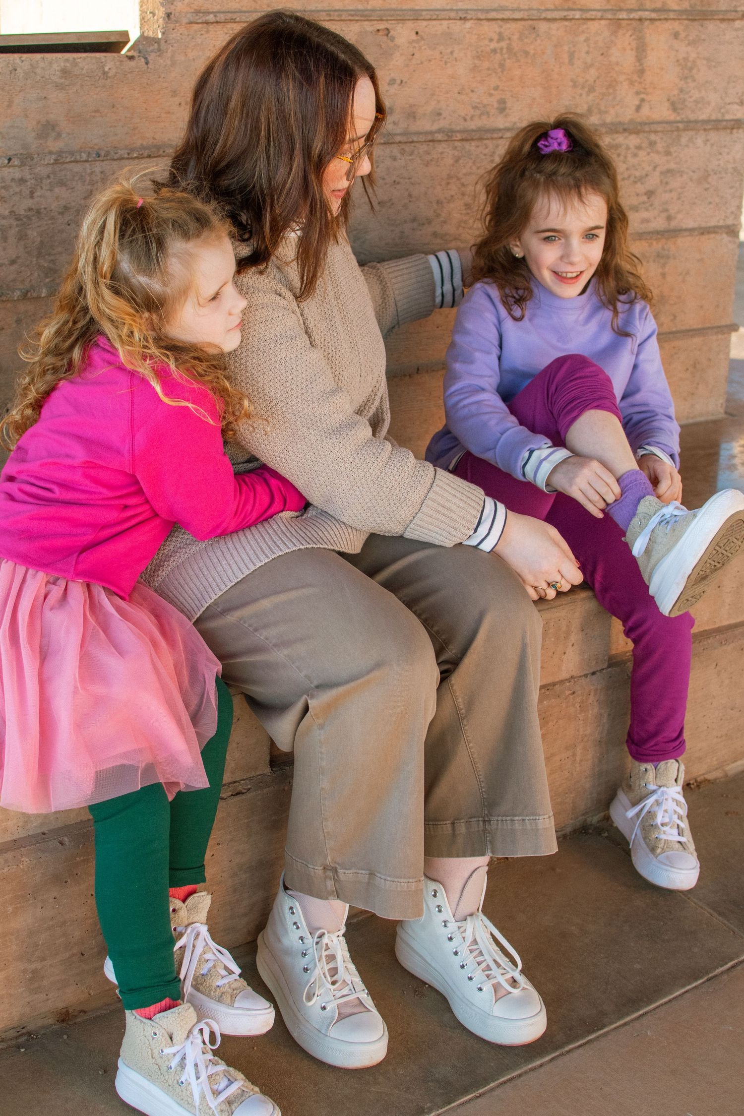 A mom and her 2 daughters seated together wearing matching white sneakers and casual attire with pink and purple outfits.