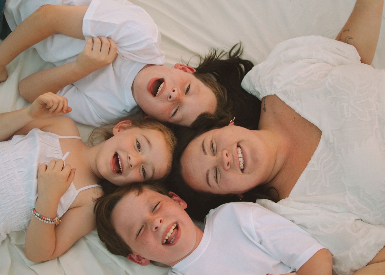 Family wearing white clothes laying together on floor with their heads in a circle formation while smiling and laughing.