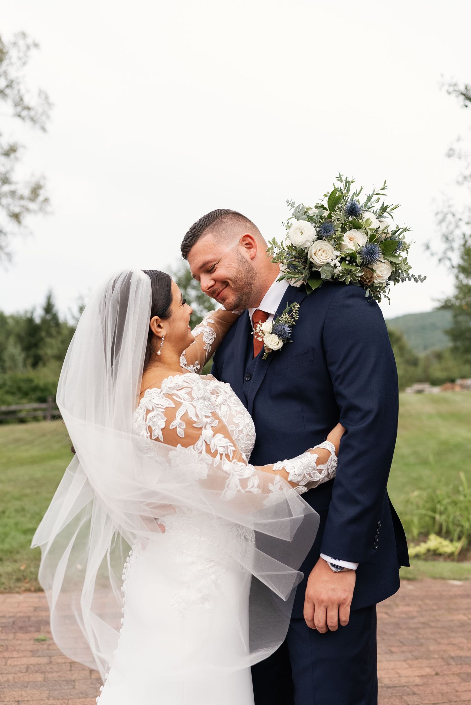Romantic wedding couple embraces outdoors with bride wearing lace dress and flowing veil.
