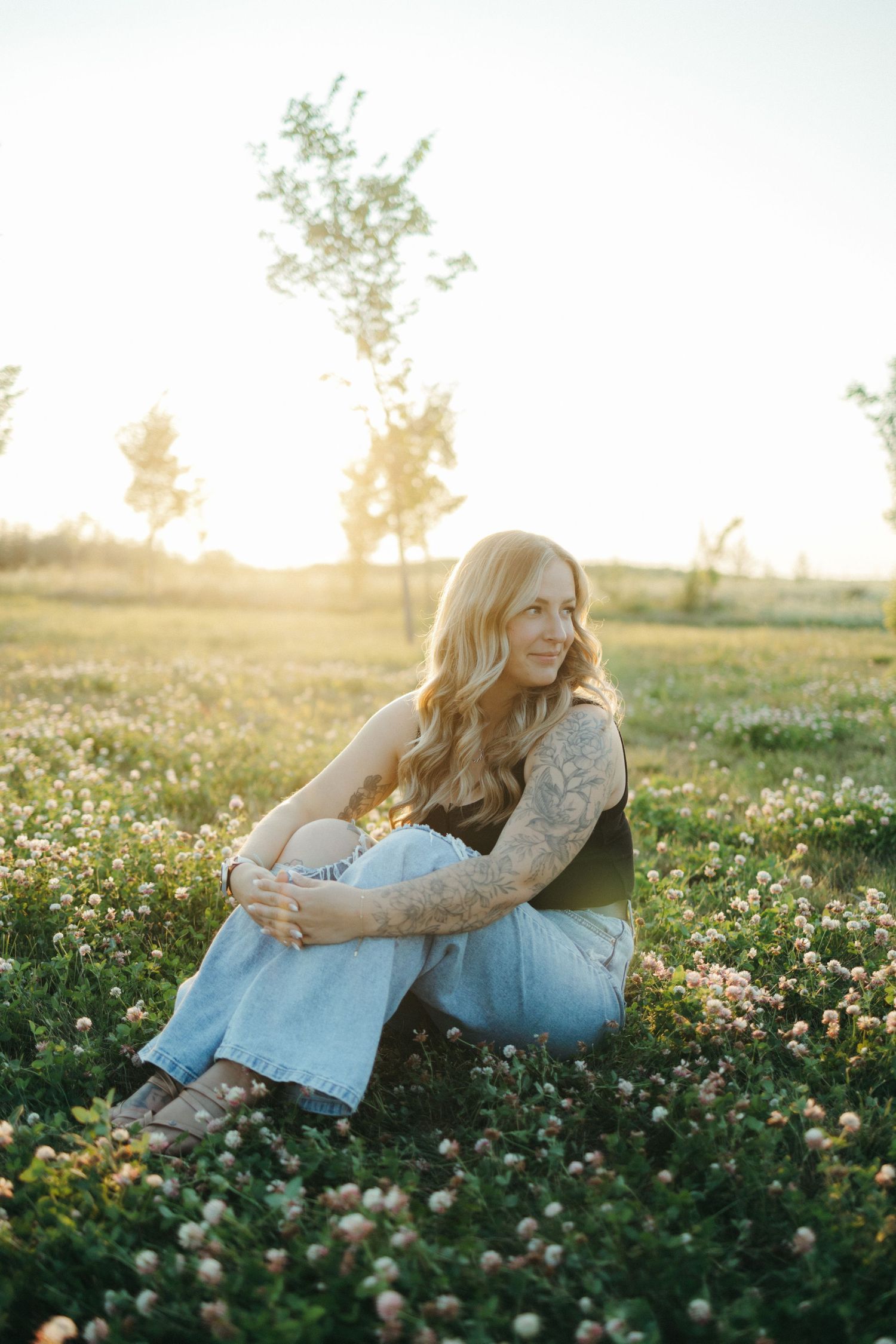 Person sitting in a field of wildflowers at sunset wearing light blue jeans and tattoos in dreamy golden light.