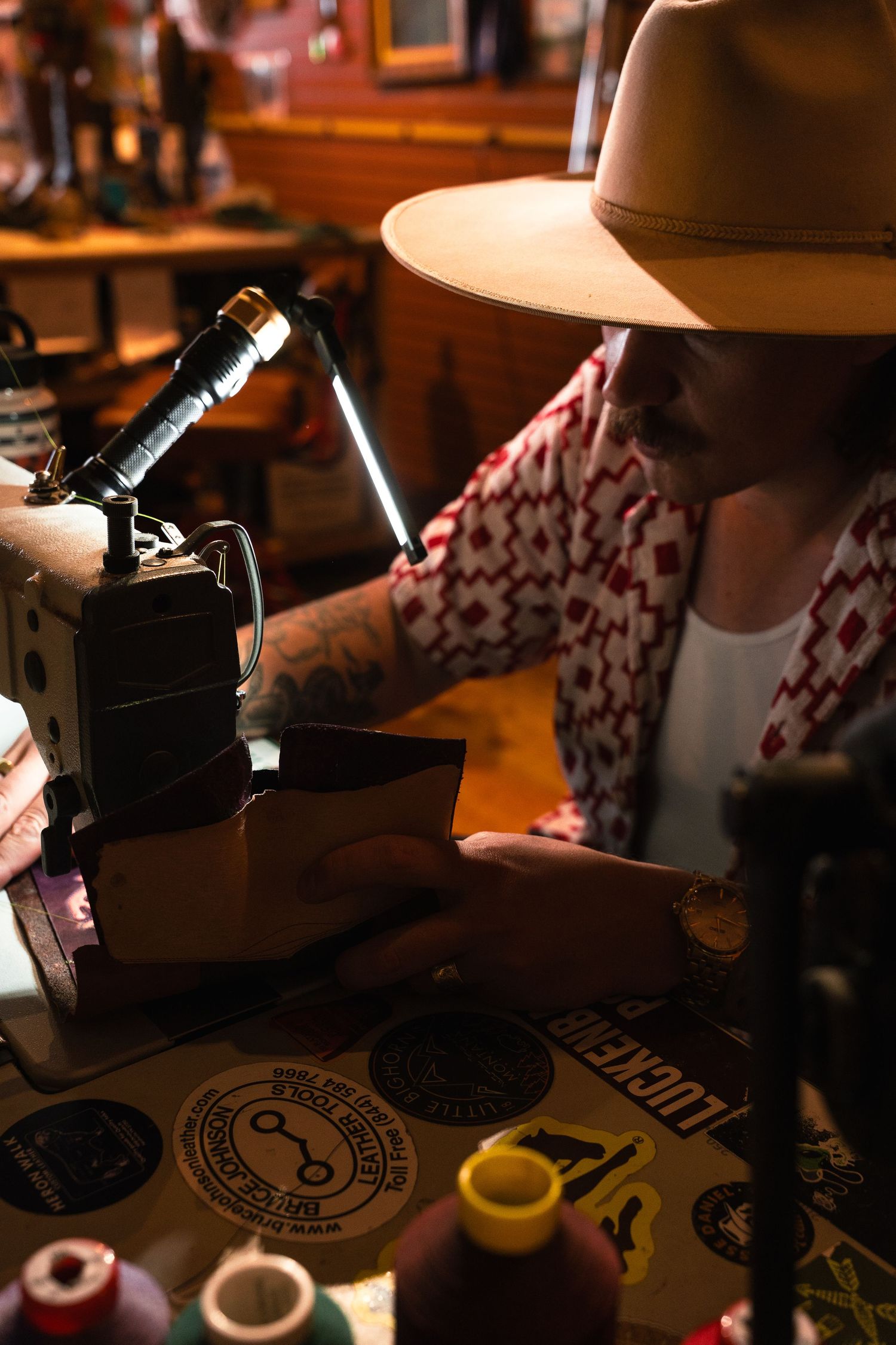 Tattooed craftsman in a wide-brim hat uses a sewing machine to stitch leather goods in a rustic workshop.