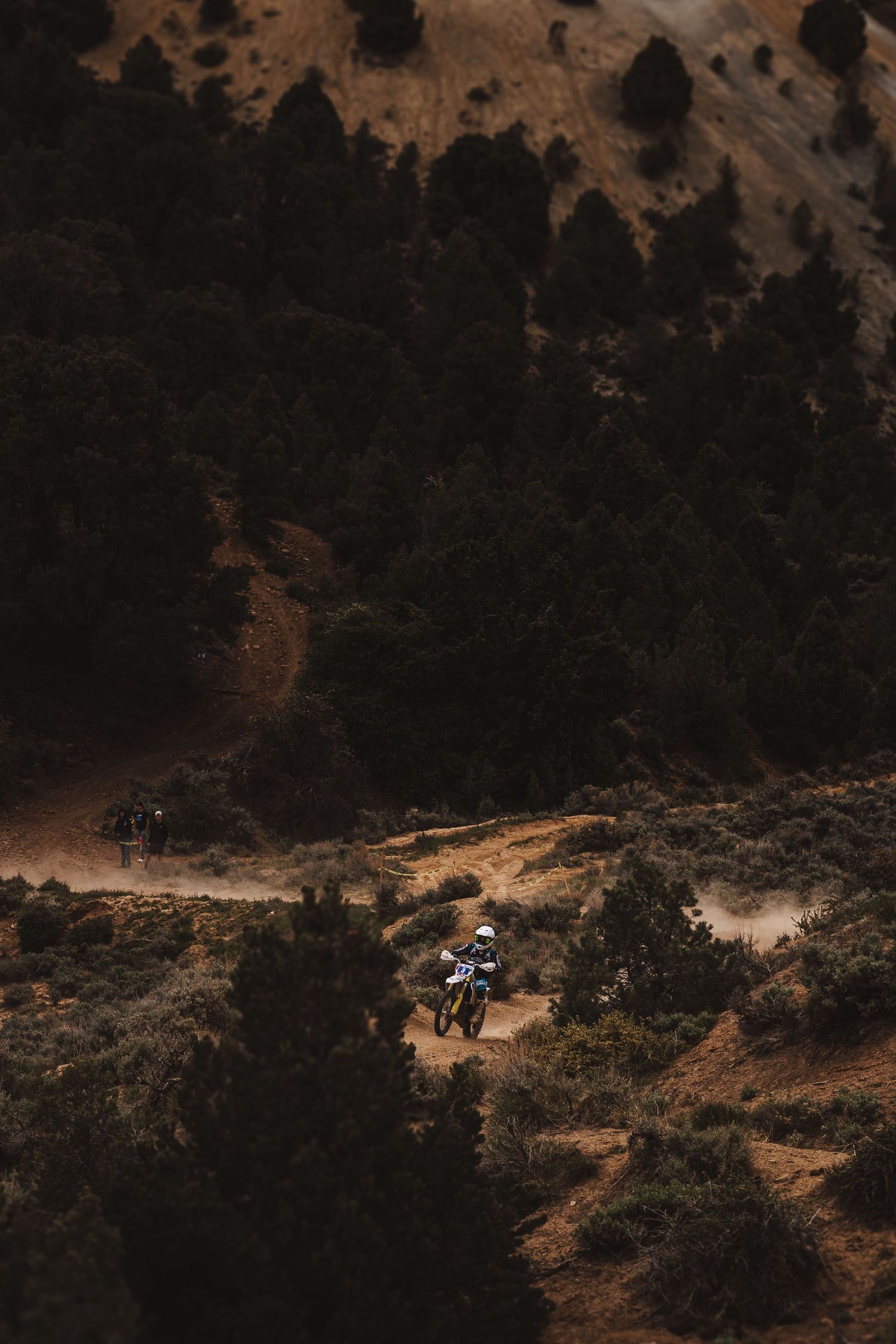 A motorcyclist rides along a dusty mountain trail surrounded by dark forests and golden hillsides in moody lighting.