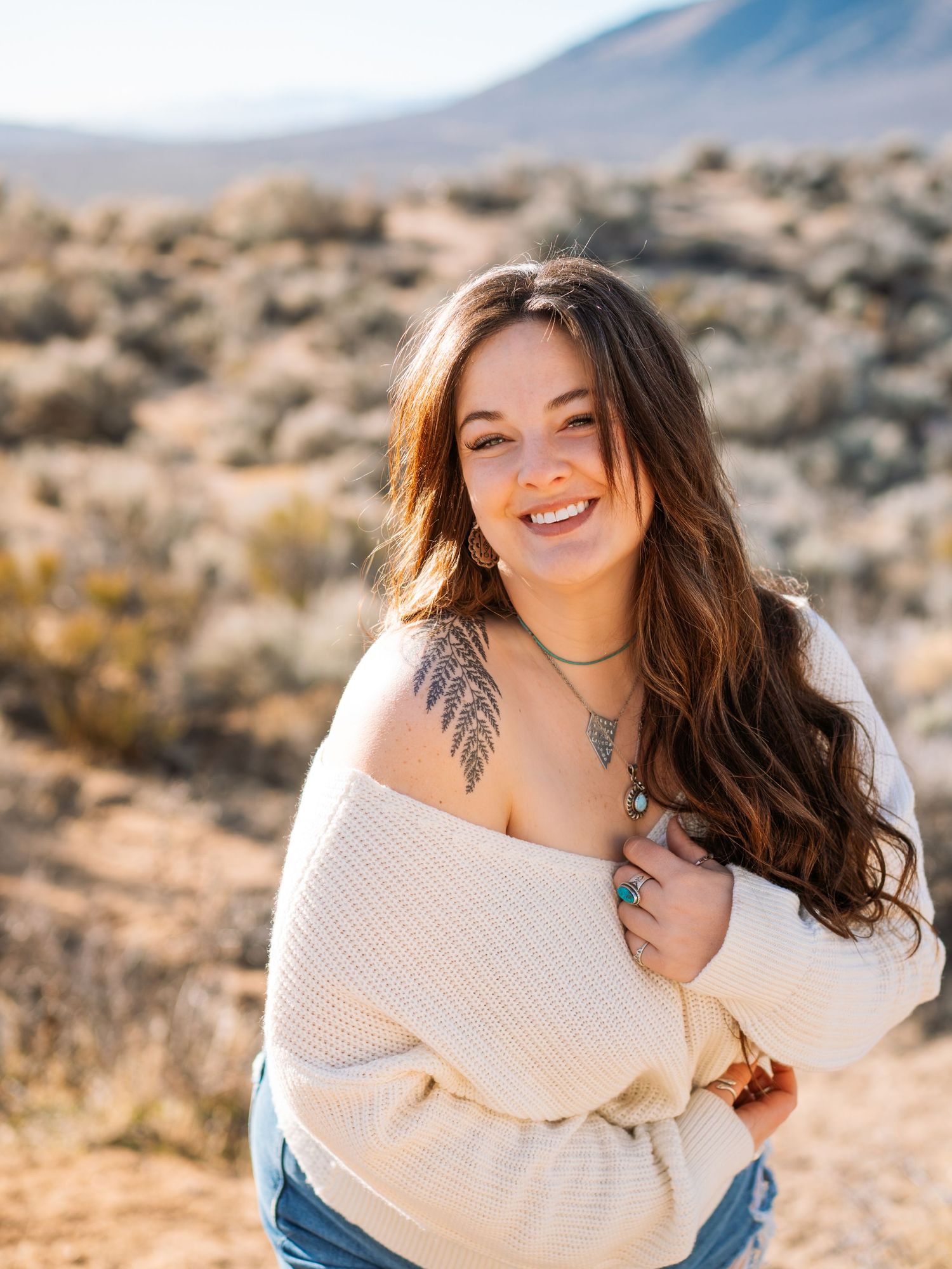 Smiling young woman with long brown hair and shoulder tattoo in cream off-shoulder sweater posing in desert landscape.