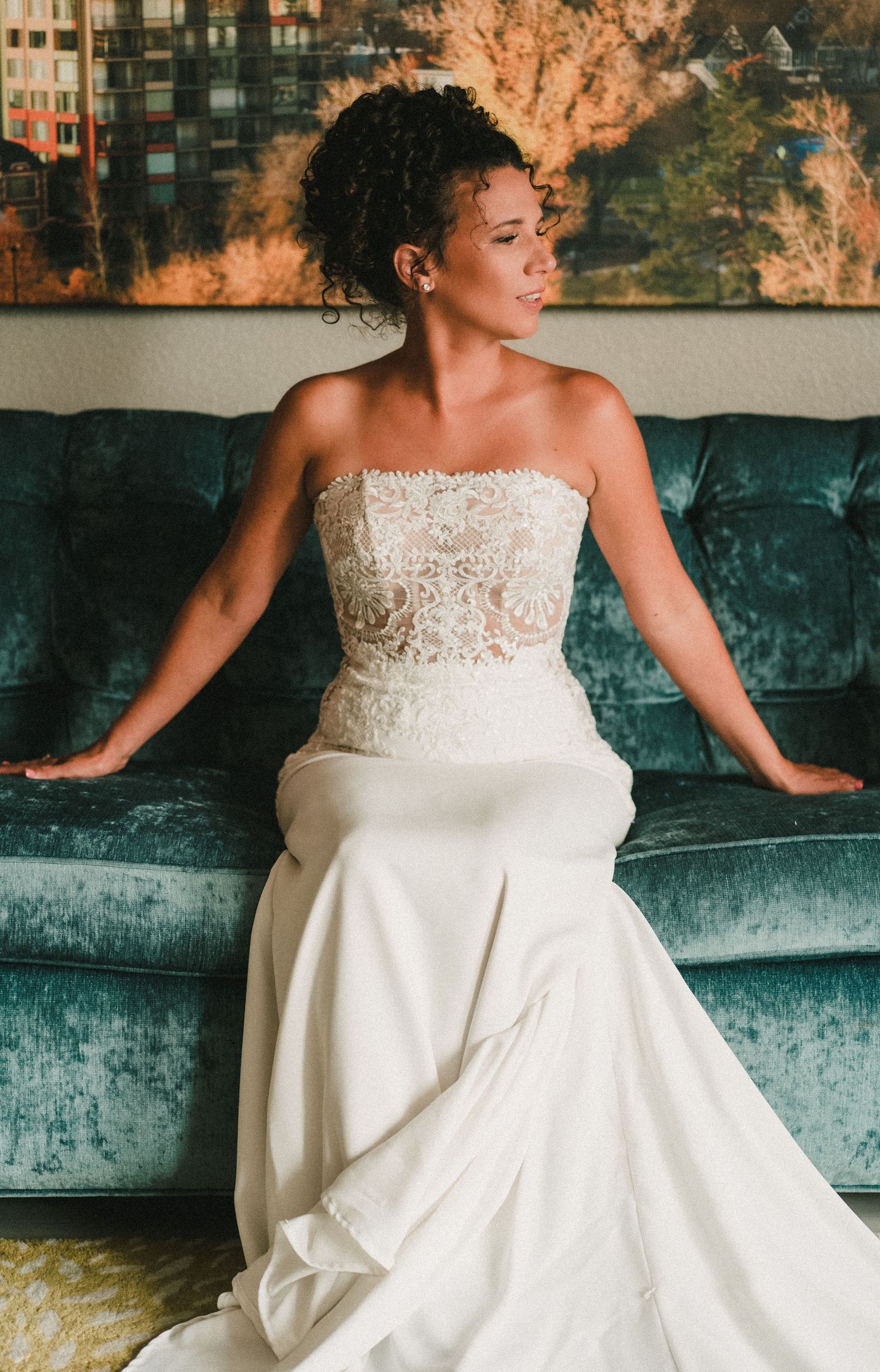 Bride in strapless lace wedding gown seated on teal velvet sofa, gazing elegantly to the side.