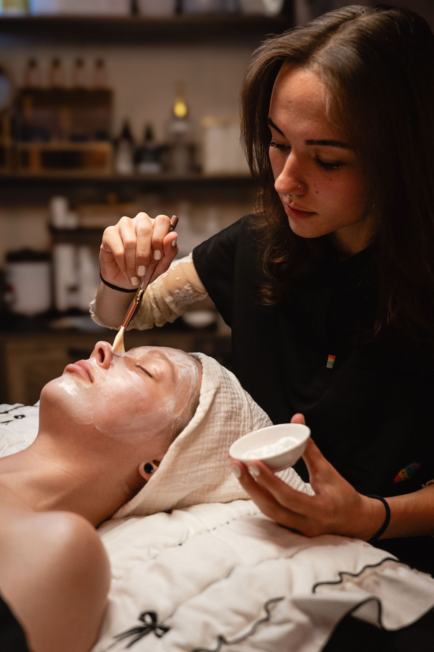 Esthetician applying a white facial mask with a brush to a client lying down in a spa salon.