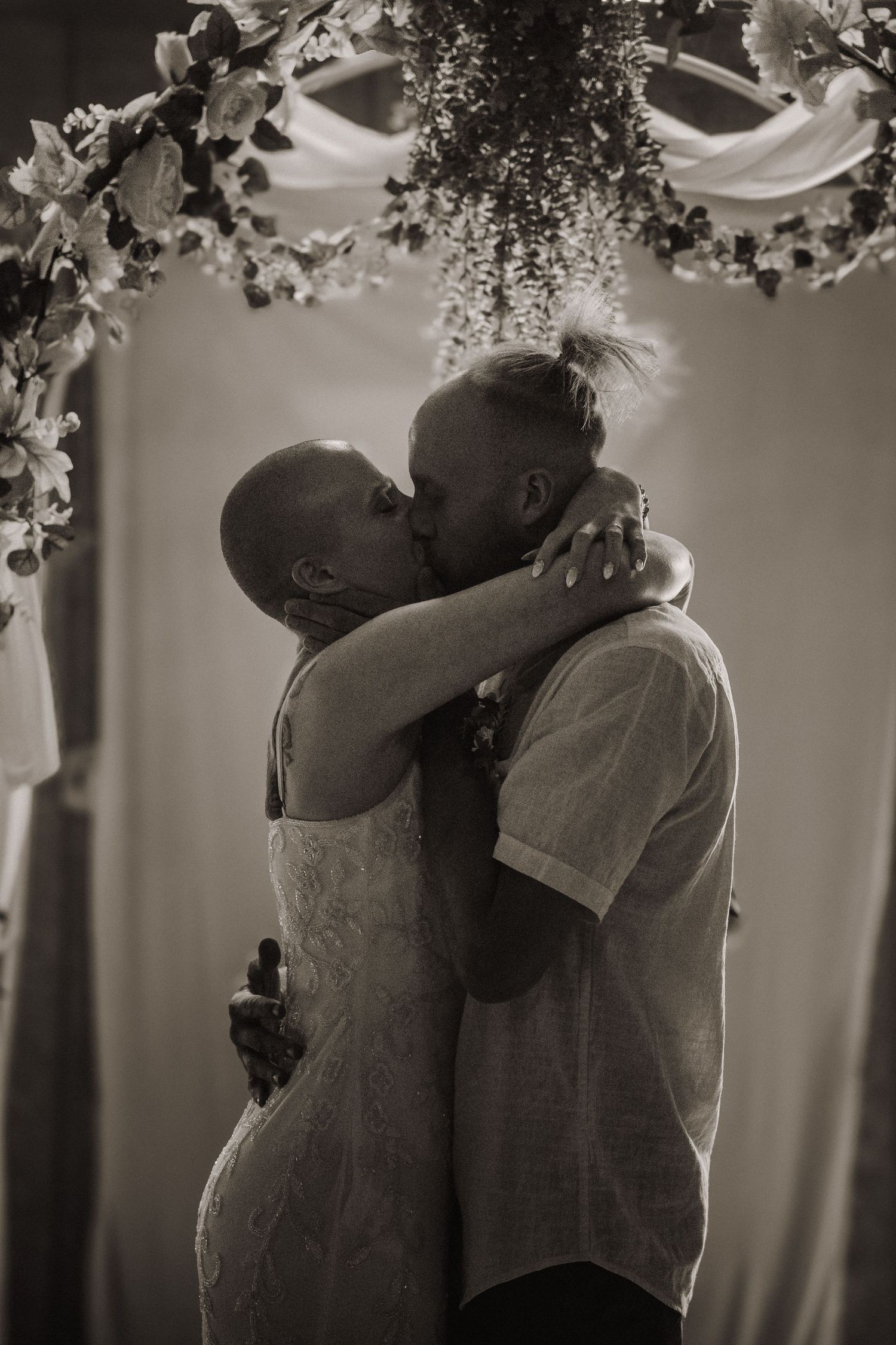 Black and white photo of a couple sharing their first kiss at a wedding ceremony beneath a floral arch.
