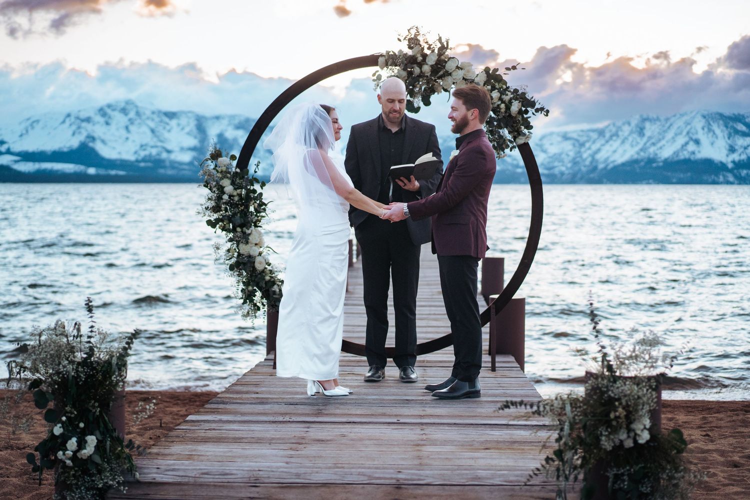 Couple exchanges vows on a lakeside dock with snowy mountains backdrop under a circular floral arch at sunset.