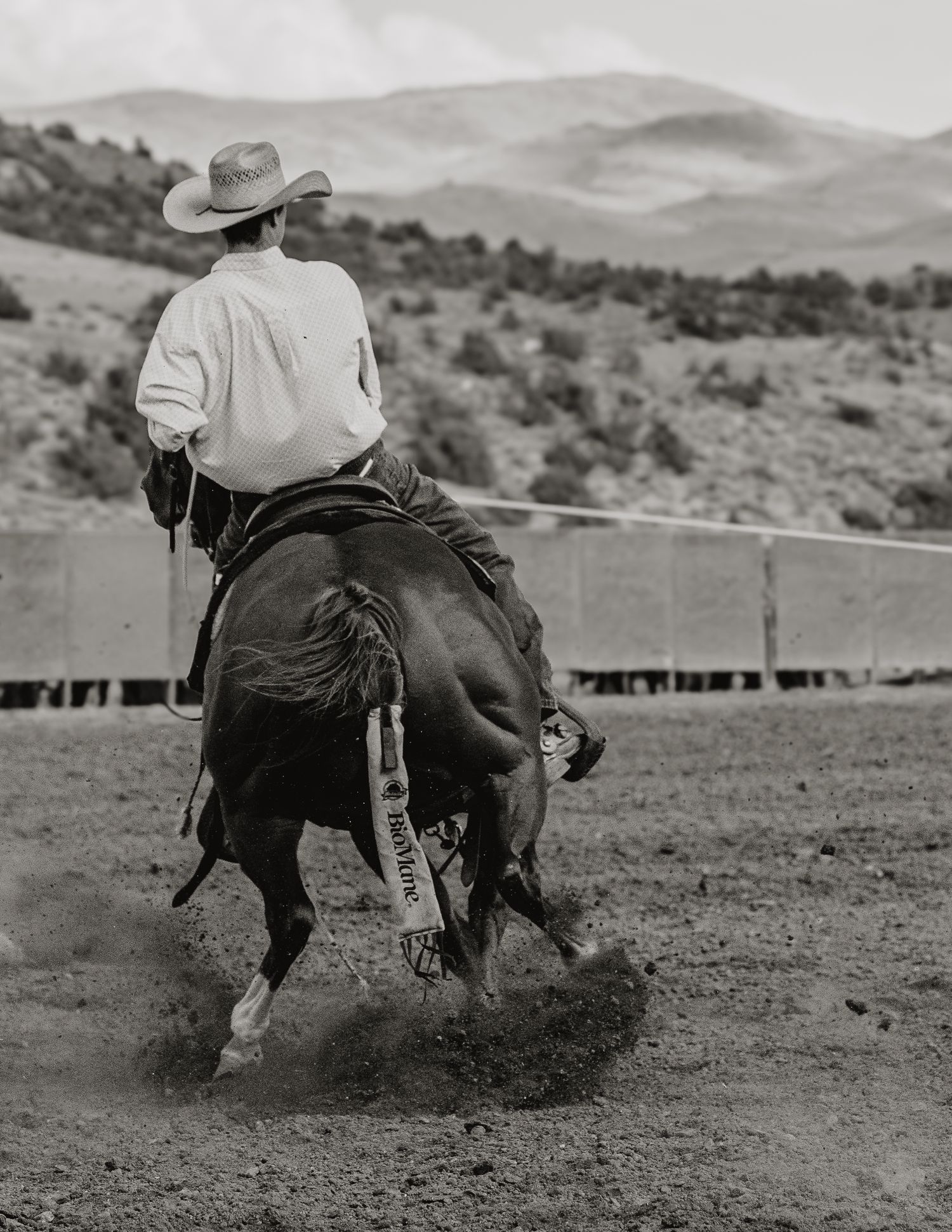 Black and white photo of a cowboy riding a horse at a rodeo with mountains in the background.