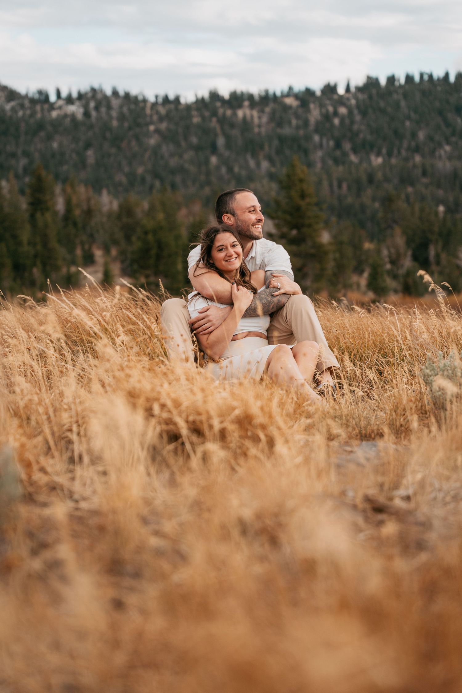 Couple embracing in golden field with pine forest backdrop, sitting together in tall dry grass during outdoor engagement session.