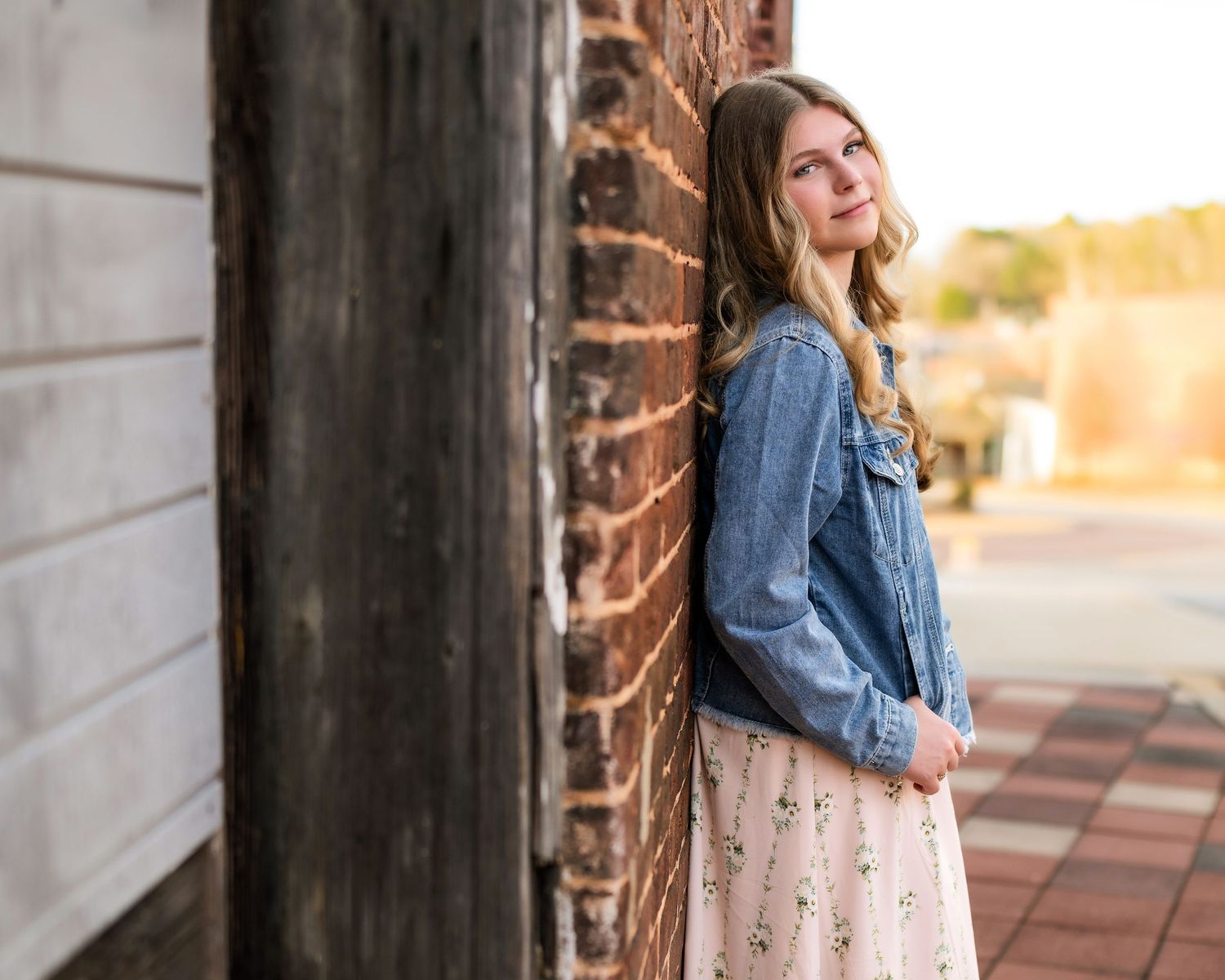 A teenage girl in a dress and jean jacket leaning up against a brick wall