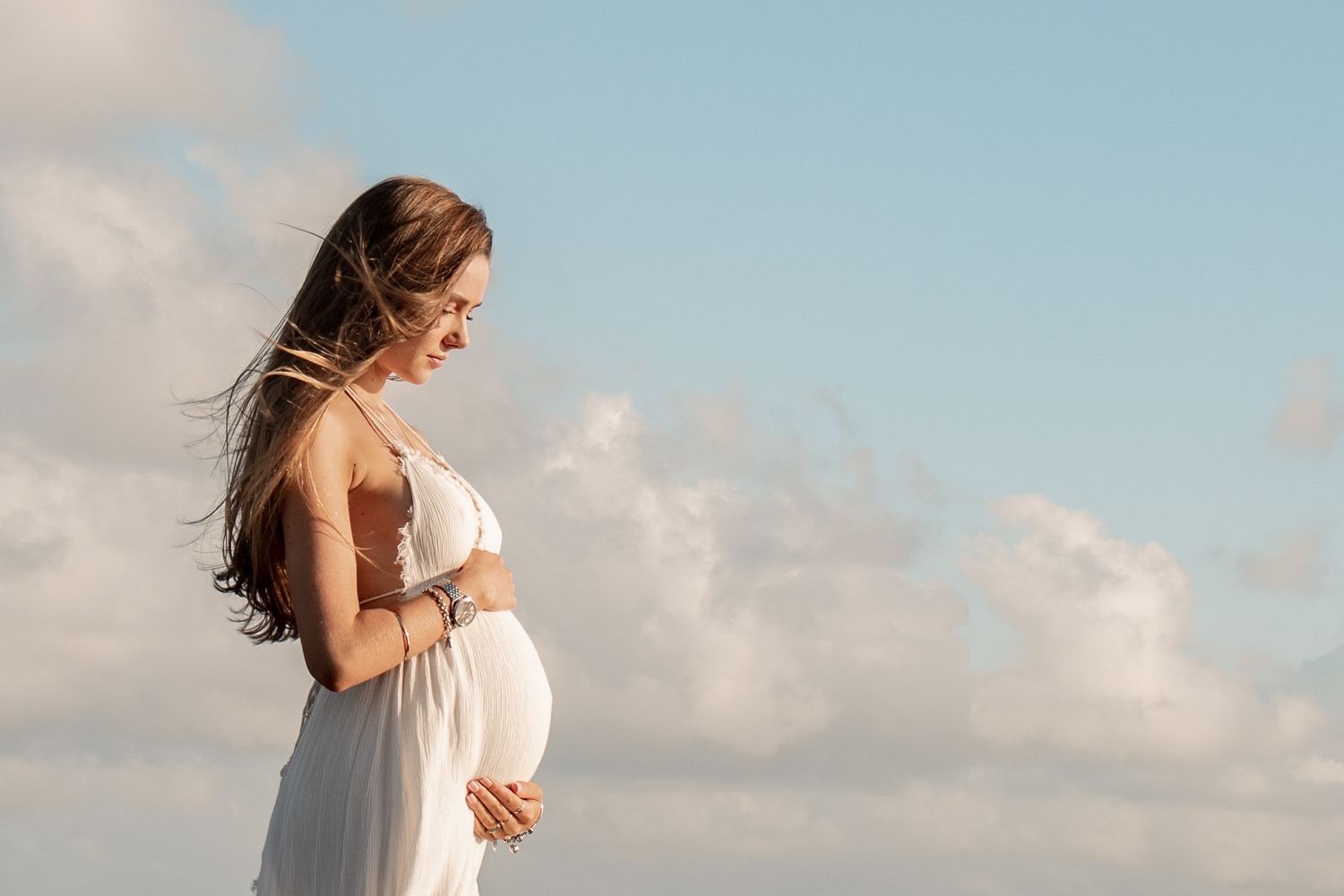 Maternity photoshoot of a pregnant woman holding her belly and looking down at her baby bump. Blue cloudy skies behind her