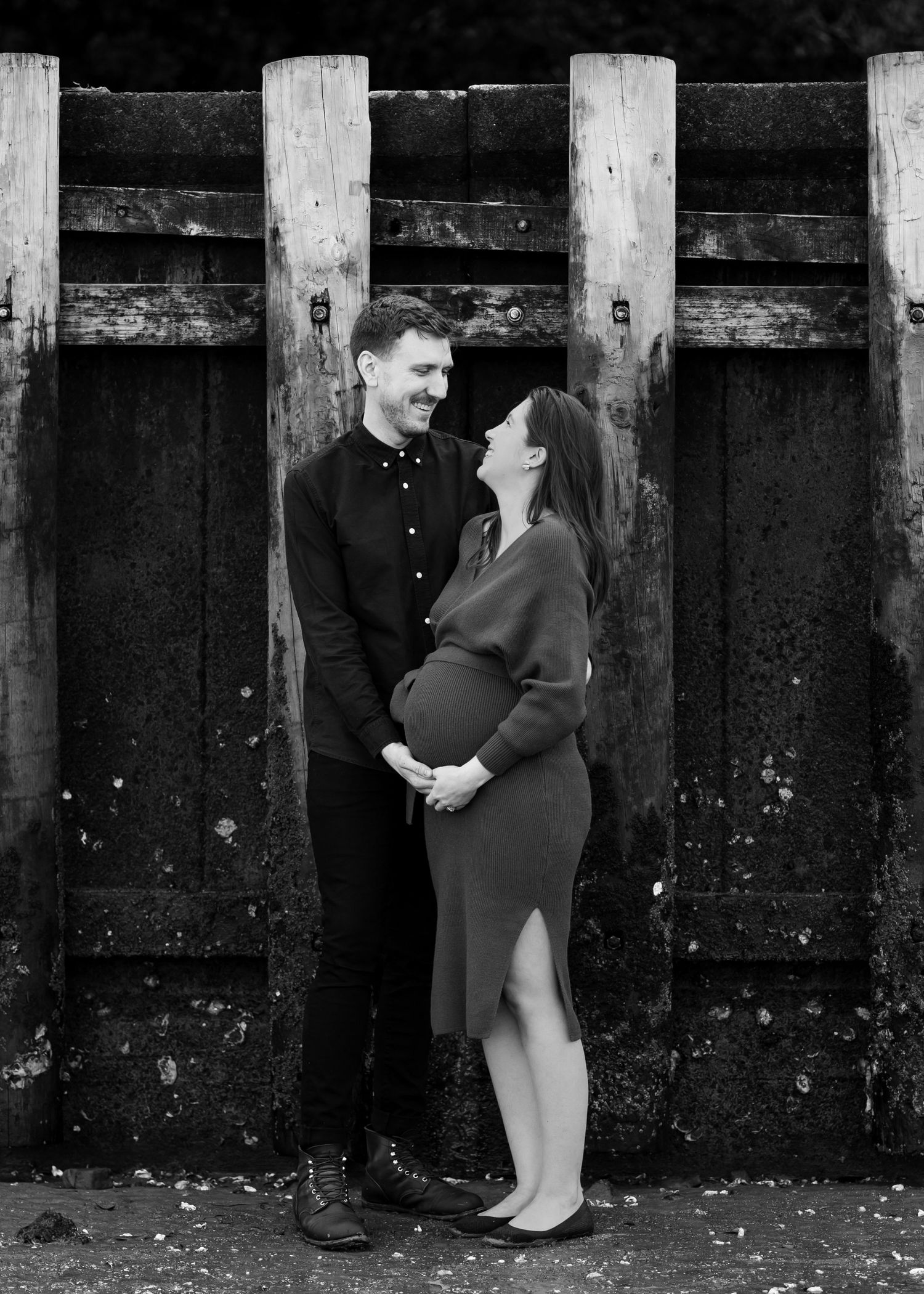 New parents to be posing at the beach for a maternity photoshoot. They are embracing, facing each other, looking into each others eyes