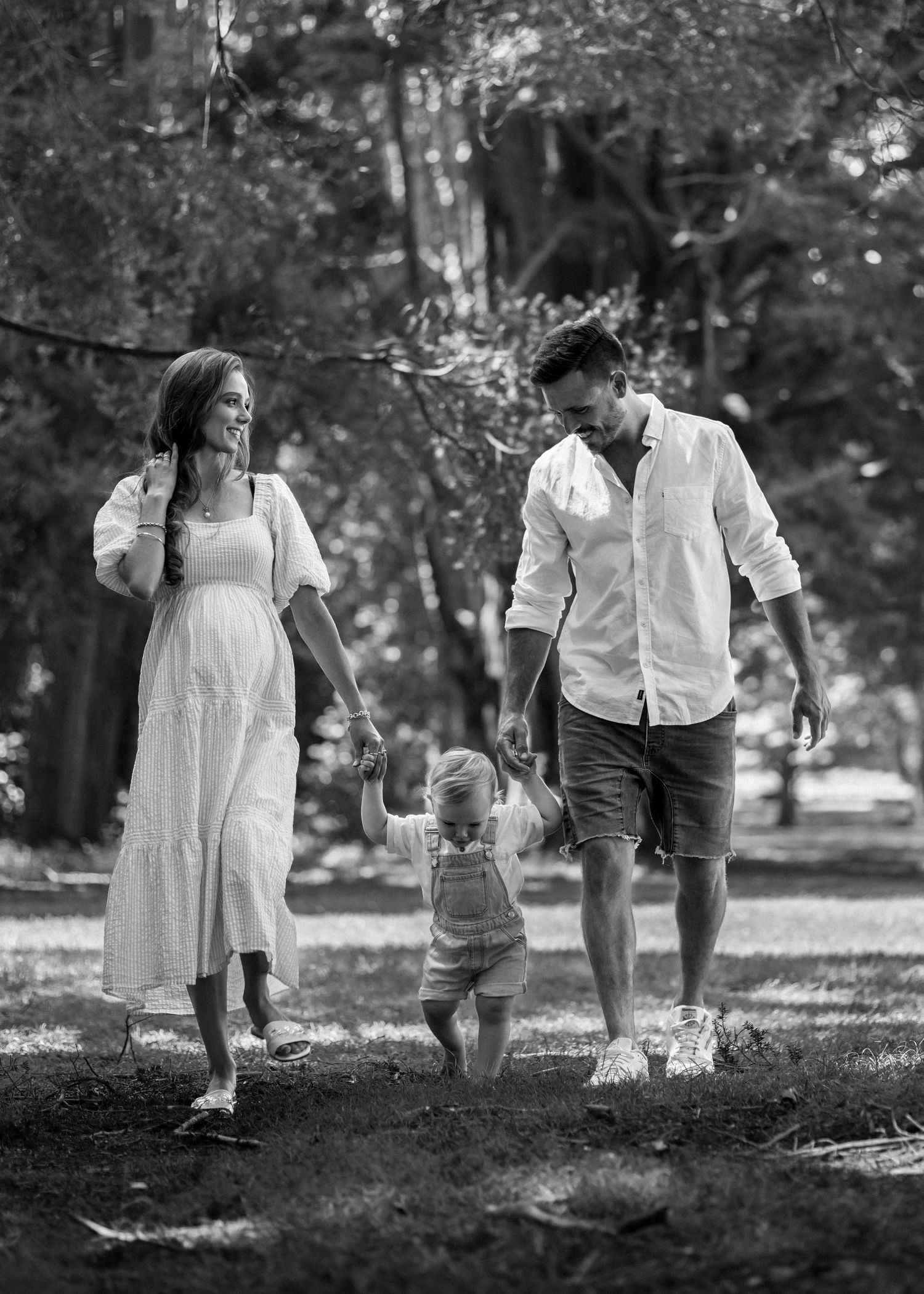 Black and white image of family of 3 holding hands walking in a line towards camera. Outdoor forest.  Maternity photoshoot CL Photography