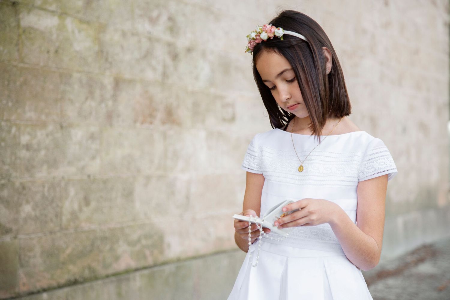 Young girl in white First Communion dress with floral headband looking down at rosary against stone wall.