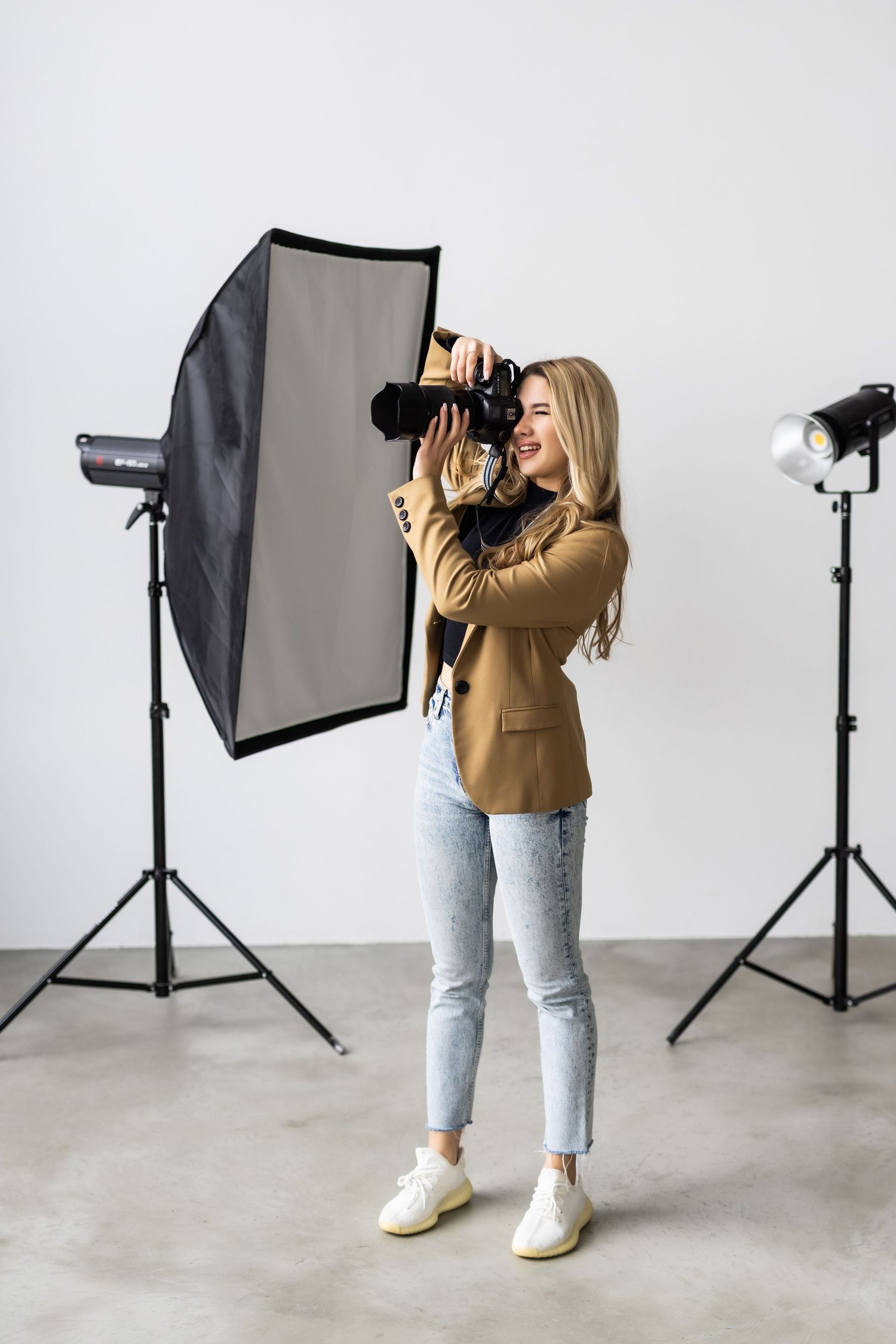 Blonde female photographer smiling while shooting with a camera in a professional studio with softbox lighting.