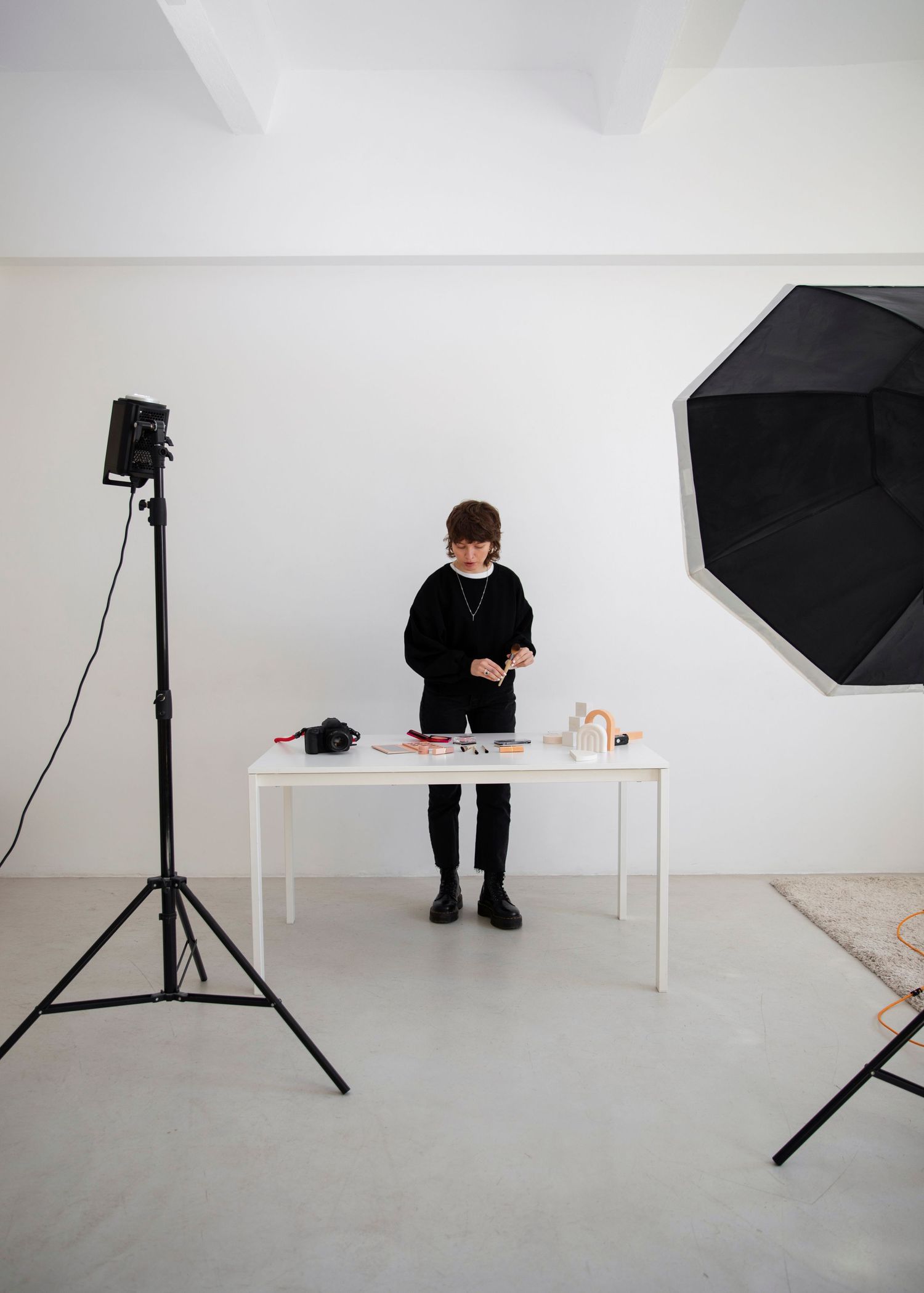 Photographer arranging items on a white table in a minimalist studio with black umbrella softbox light.