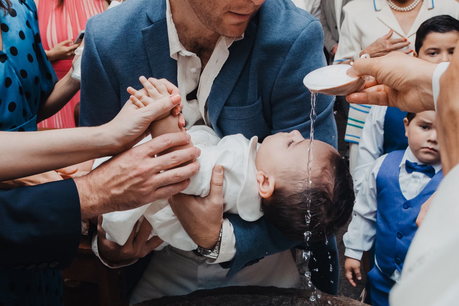 Priest baptizing infant by pouring water over head during Catholic baptism ceremony with family gathered around.