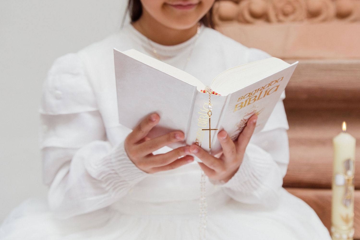 Young girl in white dress reading Bible with gold cross during First Holy Communion ceremony beside candle.