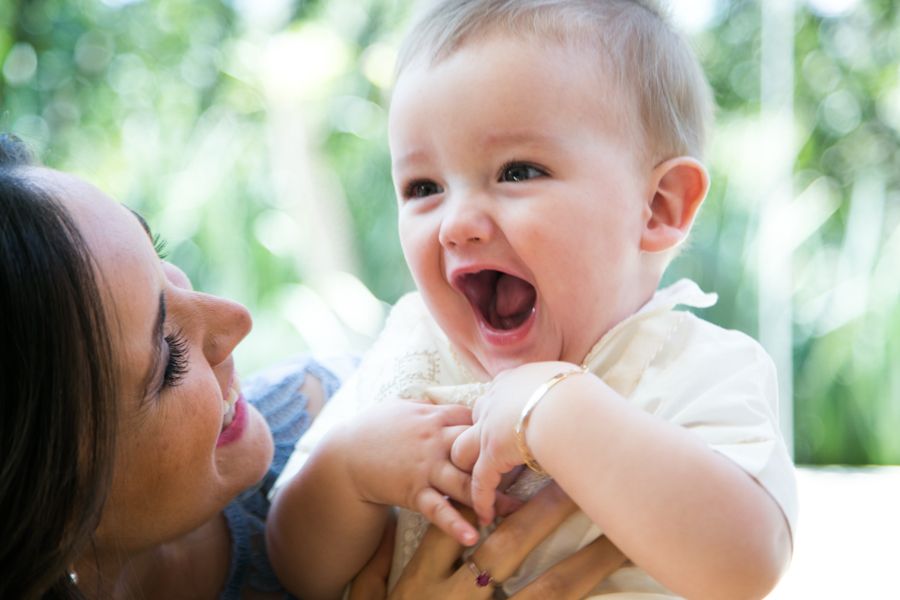 Happy laughing baby being held by smiling mother with lush green background in joyful outdoor portrait.