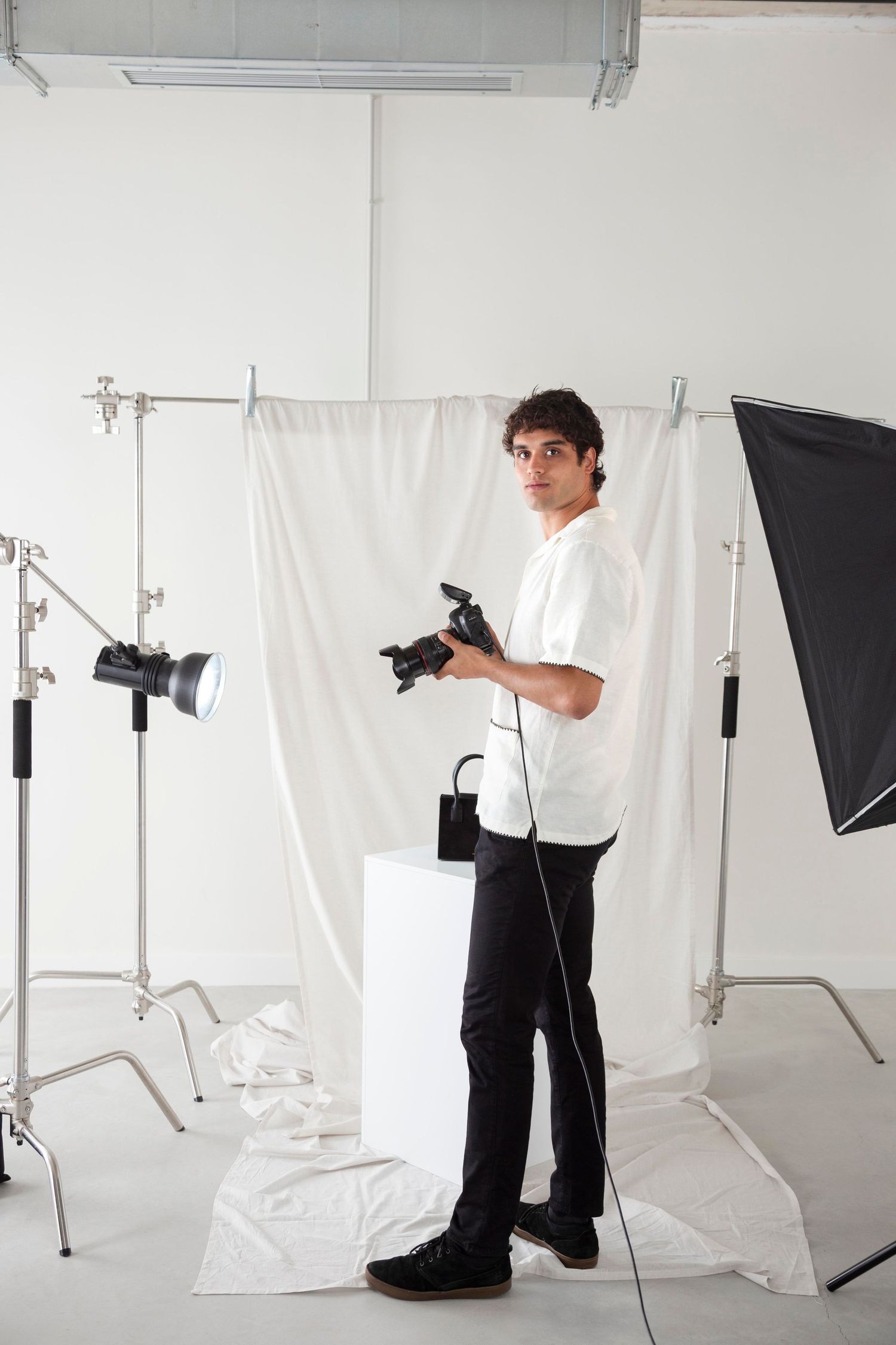 Young male photographer holding a camera in a white professional photography studio with lighting equipment.