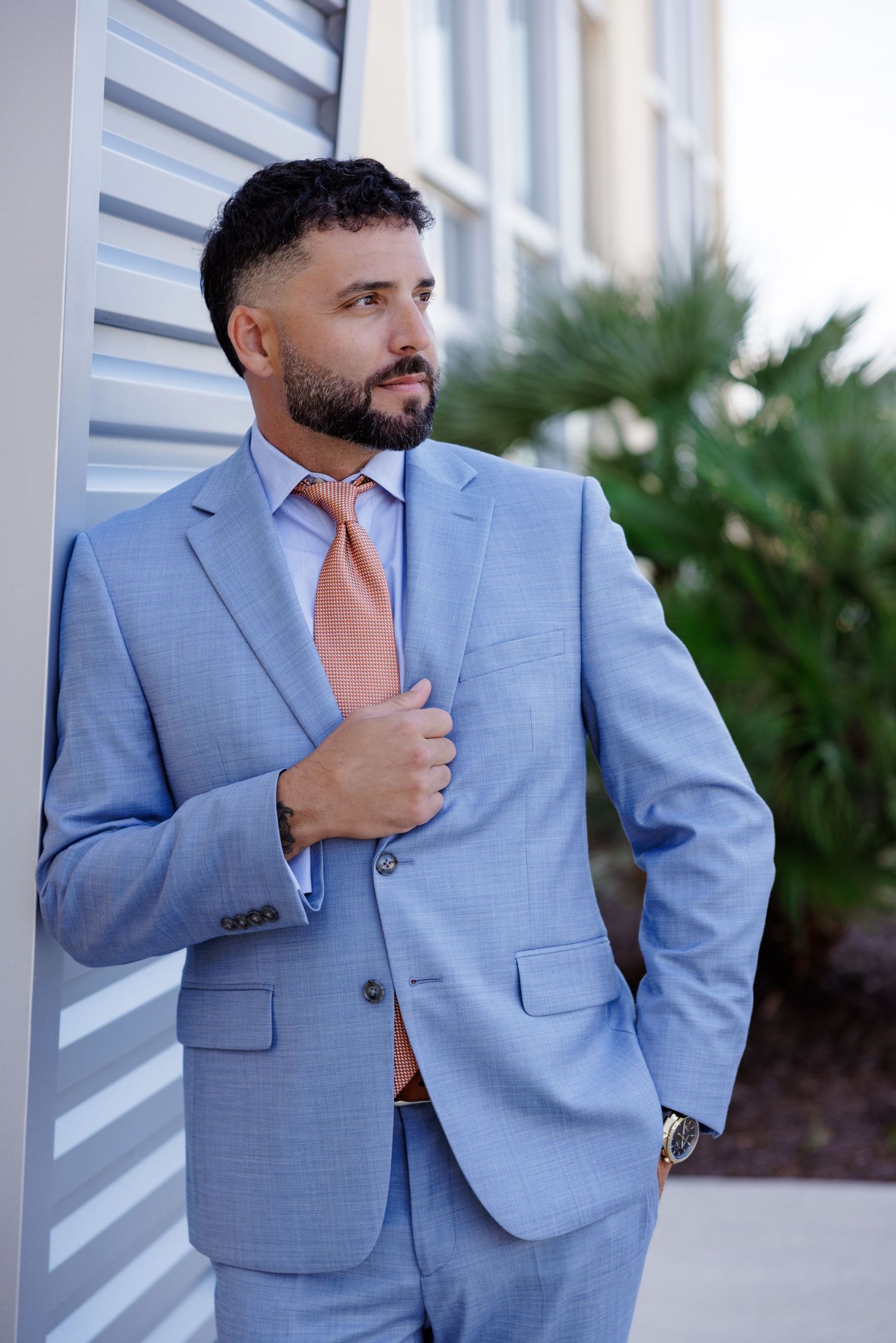 Man in light blue suit and coral tie posing against white exterior wall.